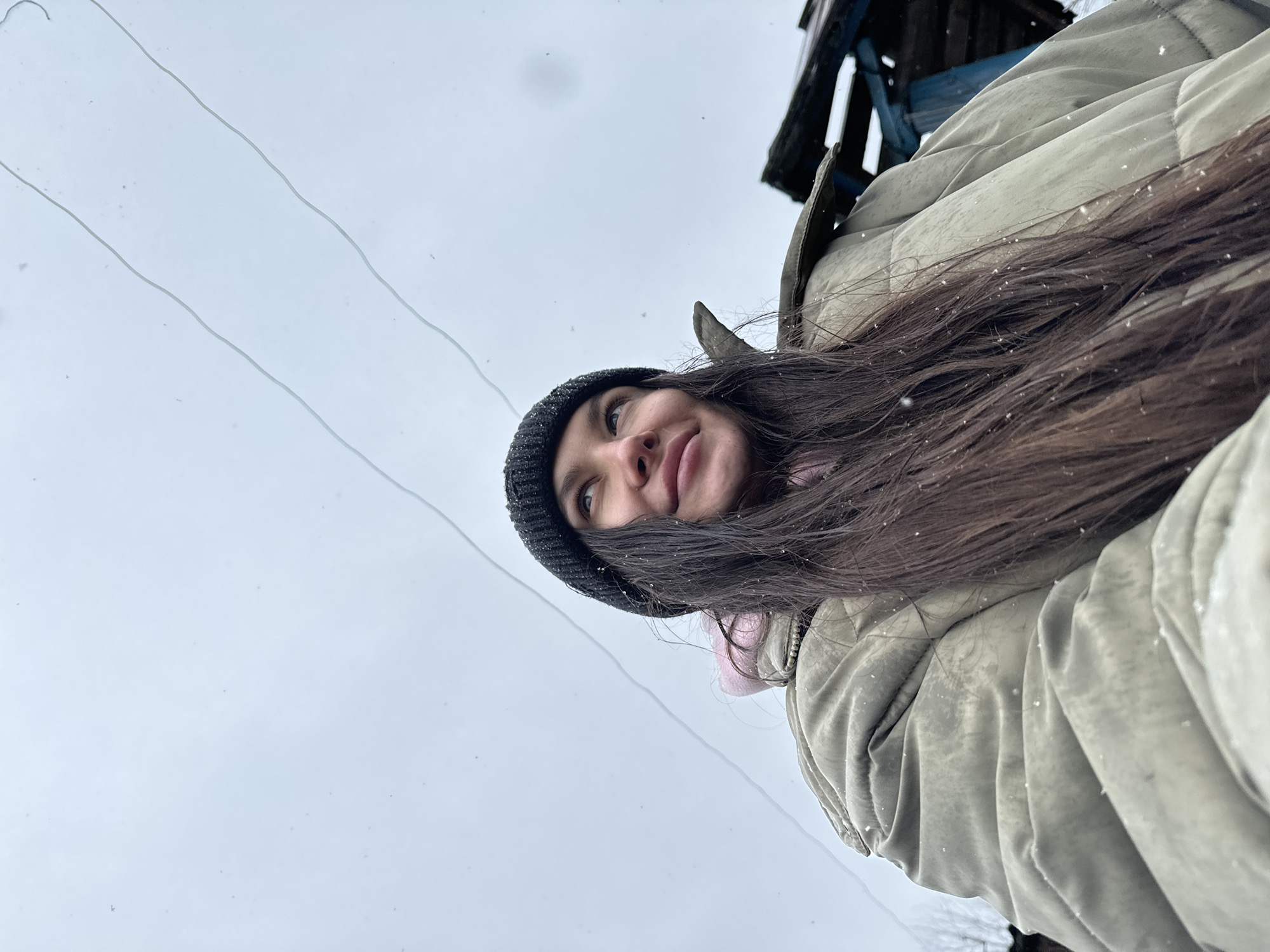 A low-angle shot depicts a woman with long, brown hair and wearing a black beanie. She is wearing a beige, quilted winter coat and a pink scarf. It is snowing, and snowflakes are visible on her hair and coat. In the background, there are power lines against a gray sky. Part of a wooden structure is visible in the upper right corner of the image. The location appears to be the city of Unknown, Unknown.