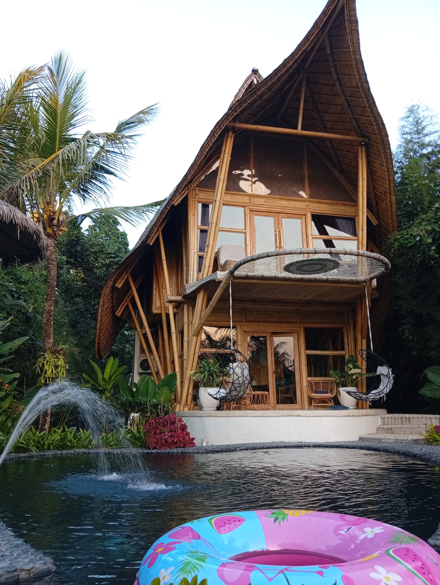 A multi-story bamboo structure with a thatched roof stands amidst lush greenery in Tampaksiring, Indonesia. The building features large glass doors and windows, several balconies, and bamboo supports. In front of the structure is a dark-colored swimming pool with a fountain creating splashes of water. A pink and blue inflatable ring with flamingo and fruit patterns floats in the foreground of the pool. Two hanging chairs are suspended from the building's second level, and rattan chairs are visible on a lower balcony. Palm trees and dense foliage surround the property. The scene appears to be a resort or vacation rental.