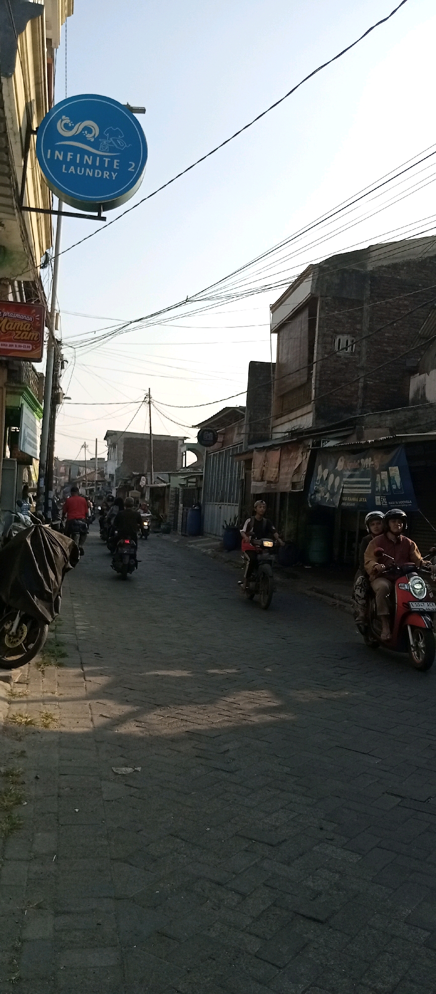 The image shows a street scene in Wadungasri, Indonesia. The street is paved with interlocking bricks and is lined with buildings on both sides. Several motorcycles are visible, with some people riding them. The lighting suggests it is daytime, possibly late afternoon, with shadows cast on the street. On the left side of the street, a circular blue sign with white text reads "INFINITE 2 LAUNDRY." Further down the street, a red motorcycle carrying two individuals is in motion. The buildings appear to be a mix of residential and commercial structures. Overhead, numerous electrical wires are strung across the street.