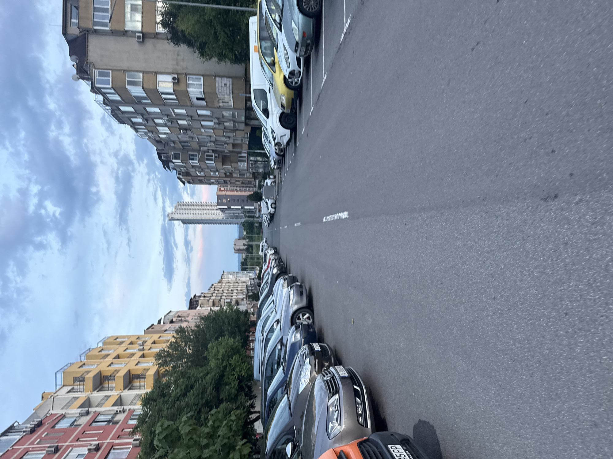 The image depicts a street scene in Burgas, Bulgaria, characterized by a row of parked cars on both sides of a paved road. On the right side of the road, multiple vehicles are parked in designated spaces, including a silver car and a white van. On the left, similar cars are parked, creating a dense row of vehicles. Buildings stand on both sides of the street, with apartment blocks and other structures of different colors and styles. The sky is visible above the buildings, showing blue patches with some cloud cover, hinting at either early morning or late afternoon light. The pavement of the street is gray and marked with white lines.