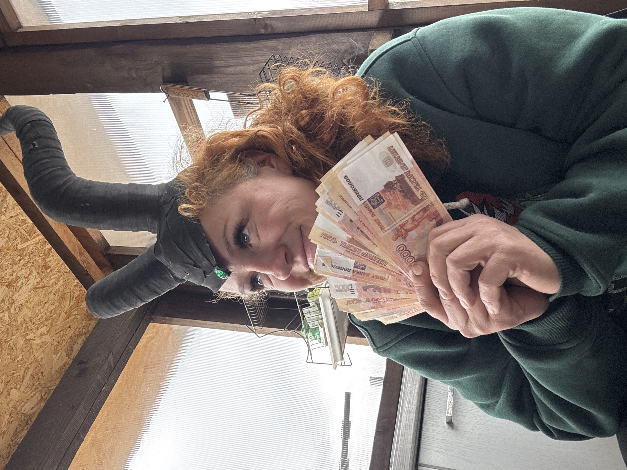 An image from Chaikovsky, Russia, features a person, likely a woman, with reddish-blonde curly hair, holding a fan of banknotes and looking directly at the camera with a pleased or confident expression. She is wearing a dark green hooded sweatshirt. A dark band or scarf with a small green embellishment is visible on her head. Her left arm, positioned to the left, is covered in a dark, possibly padded or wrapped sleeve or prosthetic, which has a textured, slightly worn appearance.
In her right hand, she holds several light-colored banknotes. Visible text on these notes, confirmed by OCR, clearly states "ПЯТЬ ТЫСЯЧ РУБЛЕЙ" (Five Thousand Rubles) and "БИЛЕТ БАНКА ПРИКОЛОВ" (Joke Banknote), indicating they are novelty items. The number "5000" is also visible on the notes.
The setting appears to be an indoor space, possibly a greenhouse or shed, characterized by wooden beams and translucent corrugated panels in the background, allowing natural daylight to enter. A metal rack is partially visible. The image is captured from a low angle and is presented rotated 90 degrees counter-clockwise, portraying a casual, lighthearted moment.