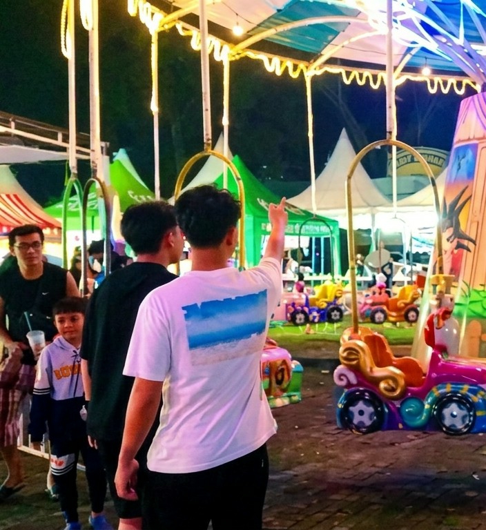 Here's a forensic description of the image:
**Location:** Medan, Indonesia, based on metadata.
**Setting:** Outdoor amusement park at night. String lights are visible overhead and illuminating the scene.
**Subjects:**
* A group of people is the focus of the image, primarily viewed from the back. These individuals appear to be in their teens or early twenties.
* A man wearing glasses and a black tank top is seen on the left side of the frame.
* A child holding a beverage is also on the left.
**Objects:**
* A carousel is visible with colorful ride vehicles.
* The carousel structure has supports, hanging seats, and a decorative roof.
* Concession stands are visible in the background.
**Actions:**
* One person in the group has their arm raised, with their index finger extended upwards.
* The people are standing and interacting within the amusement park.
**Interactions:**
* The group appears to be observing or pointing toward something in the amusement park.
**Environment:**
* The environment is illuminated by string lights.
* Tents are visible in the background, suggesting a festive atmosphere.