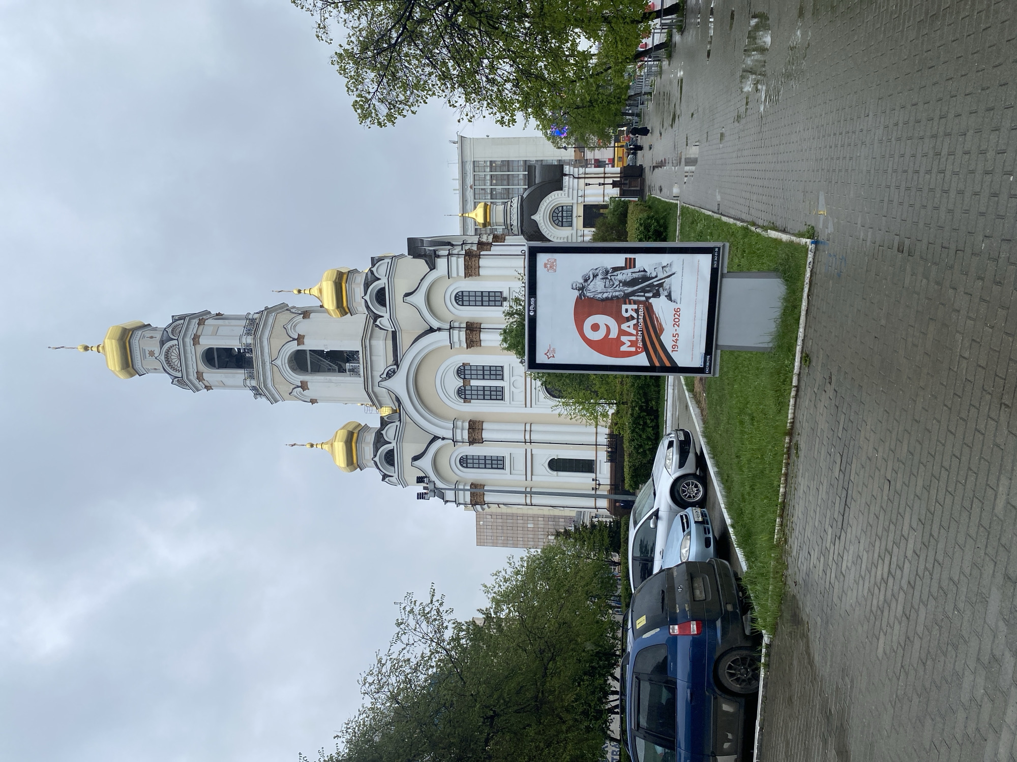 The image depicts a street scene in Yekaterinburg, Russia. In the background is a white and gold Orthodox church with a tall bell tower. A large billboard stands prominently on the street, displaying the text "9 мая" (May 9th), likely advertising Victory Day celebrations. Several cars are parked along the curb. The pavement is wet, suggesting recent rain. Barely visible figures are walking on the sidewalk, further down the road. The scene is overcast.