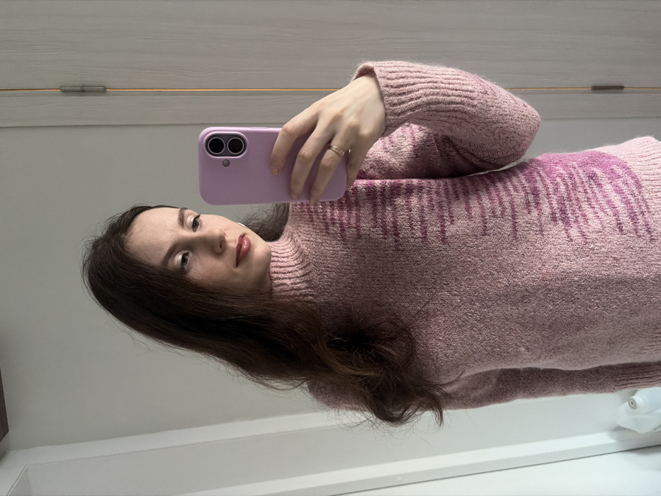 A woman in a pink and purple sweater takes a selfie in a mirror. She holds a lavender phone, the camera side facing forward. She has long brown hair and is wearing a ring on her ring finger. A small portion of a white wall and door are visible behind her.