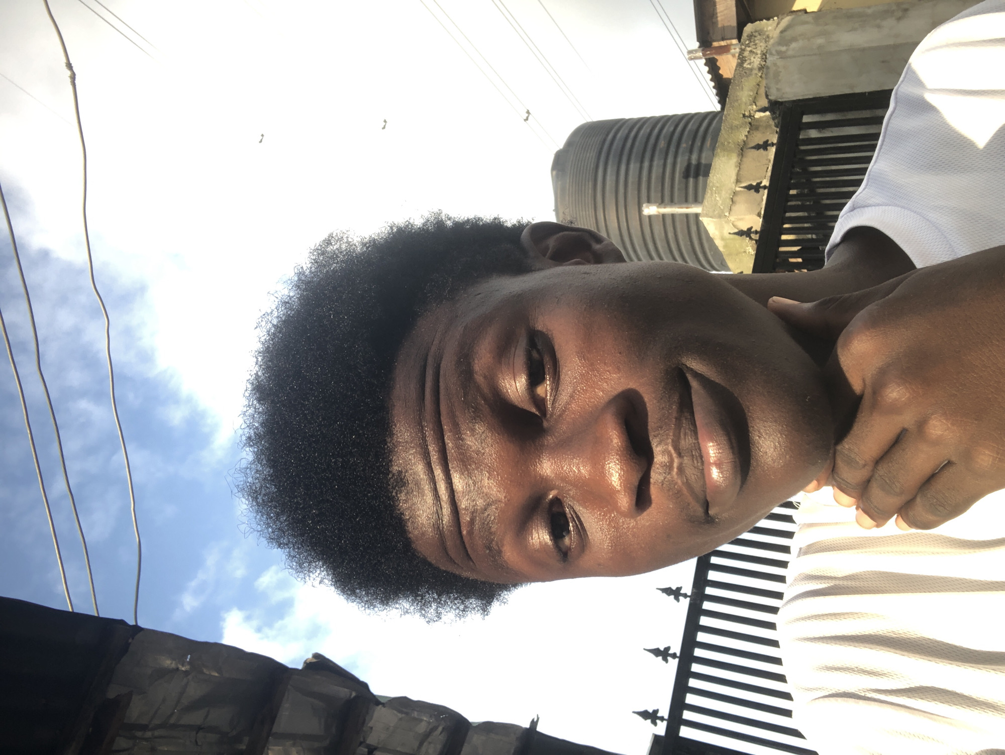 This is a self-portrait taken outdoors, likely in Ado, Nigeria. A young Black male with dark, short, curly hair is positioned in the foreground, looking towards the camera. He is wearing a white t-shirt with a ribbed texture. His right hand is raised to his chin, and his thumb and index finger are resting on his jawline.
The background consists of a bright sky with scattered clouds, suggesting daytime. Several wires are visible crisscrossing the upper portion of the image. To the left, a dark, textured structure, possibly a building facade, is partially visible. In the mid-ground, a black, ribbed water tank stands prominently. Behind the tank, there is a black metal fence with pointed finials. The overall setting appears to be an urban or semi-urban environment.
