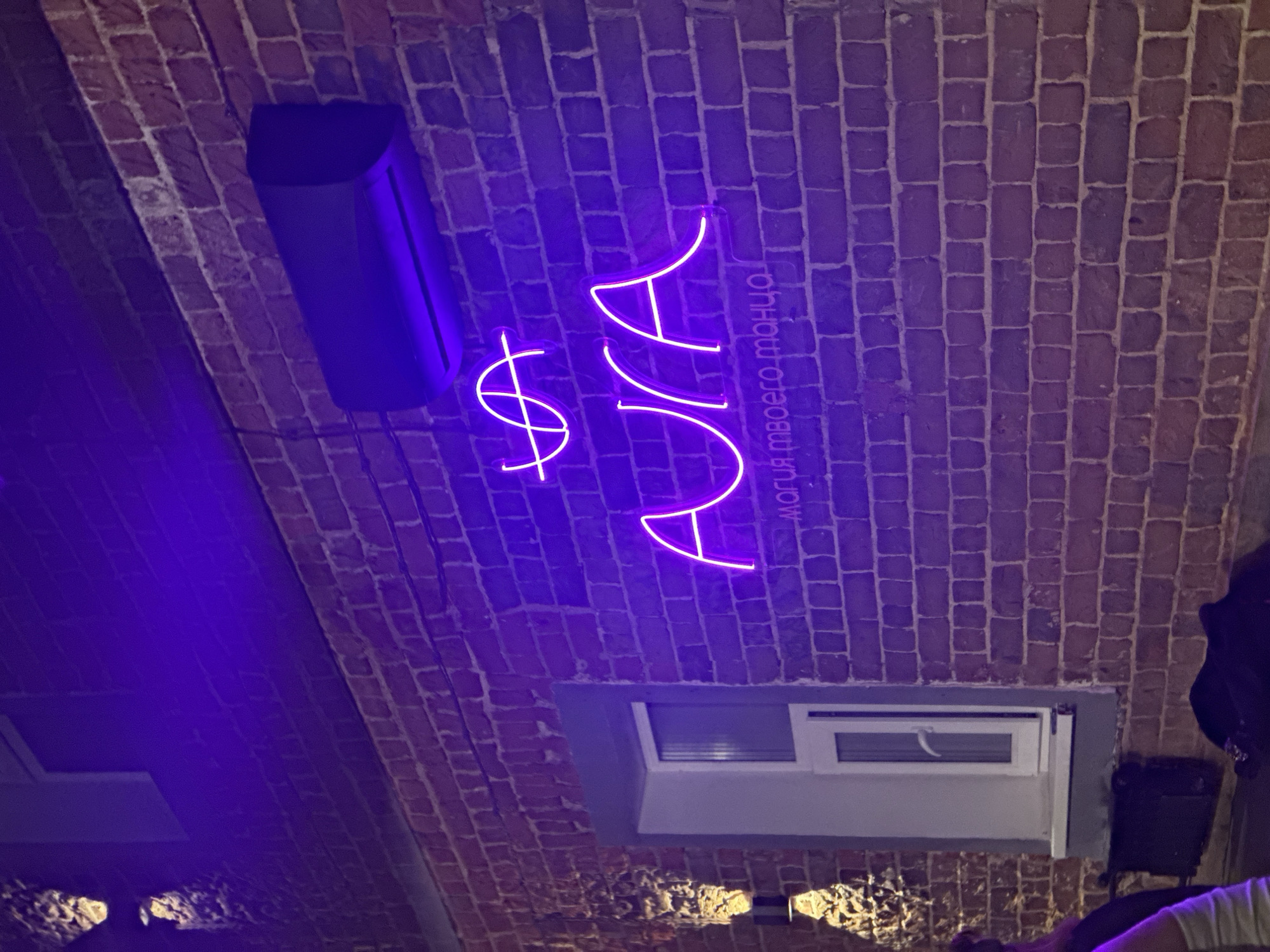 The image depicts a low-angle view of a brick wall or ceiling, possibly within an indoor space in Kazan, Russia. The wall is illuminated with a purple neon sign reading "AURA" alongside a dollar sign and a Russian phrase "магия твоего танца". Attached to the wall are a black speaker and a window or ventilation unit. Parts of other objects, including what appears to be a person wearing a light-colored garment, are visible in the lower portion of the frame. The overall ambiance appears to be dimly lit and possibly related to a nightclub or entertainment venue.