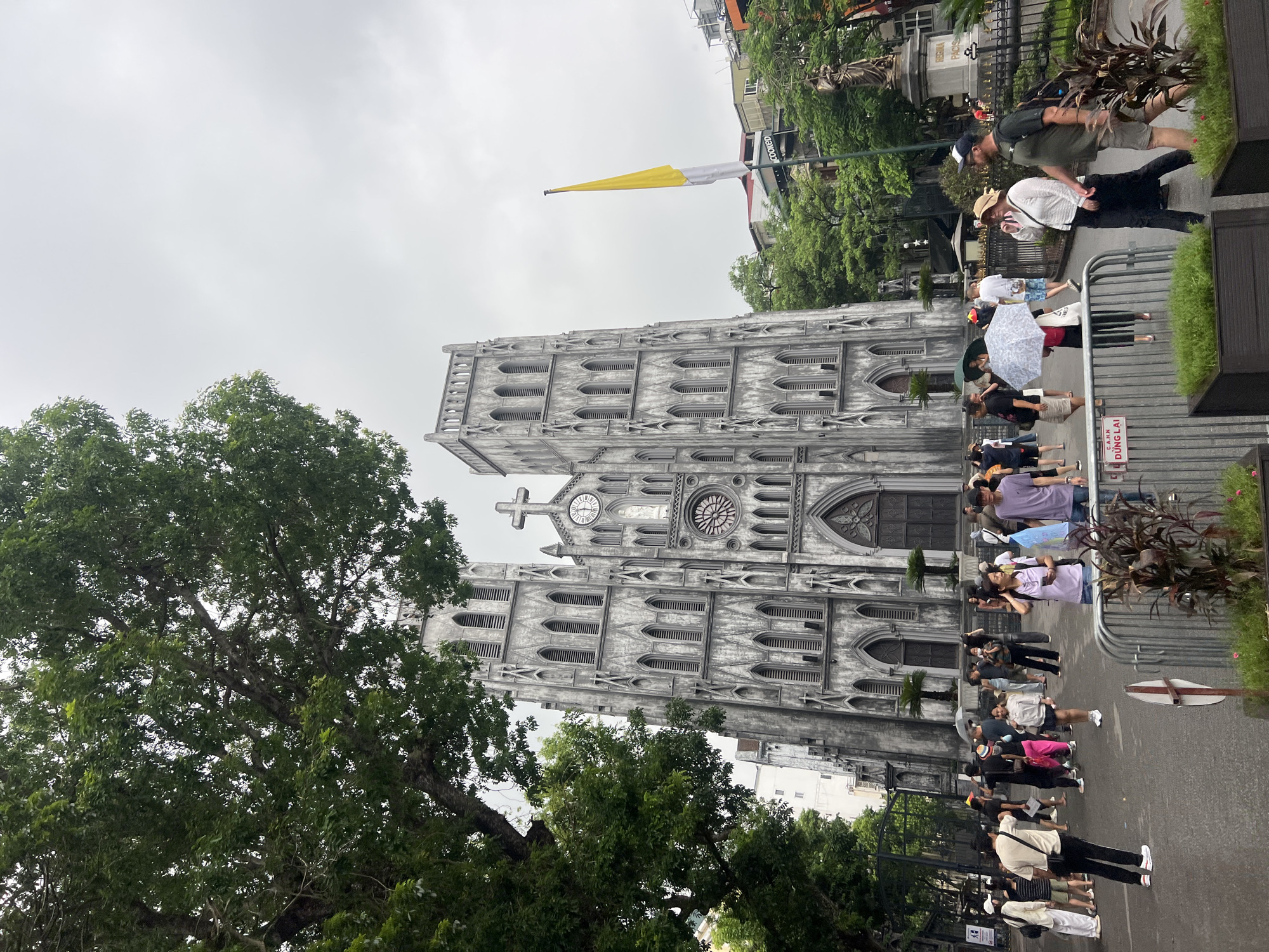 The image captures a view of St. Joseph's Cathedral in Hanoi, Vietnam, on an overcast day. The imposing Gothic Revival cathedral, constructed from stone, dominates the frame with its twin towers and ornate facade. A large tree with lush green foliage partially obscures the left side of the cathedral. In front of the building, a crowd of people, appearing to be tourists and locals, mill about. Some are holding umbrellas, suggesting light rain or protection from the sun. A yellow and white flag is visible on a pole to the right of the cathedral, alongside a statue on a pedestal. The ground in the foreground is paved, and metal barriers with planters containing greenery are present. People are dressed in casual attire, with a variety of bags and accessories. The overall atmosphere suggests a public gathering or tourist attraction in a city environment.