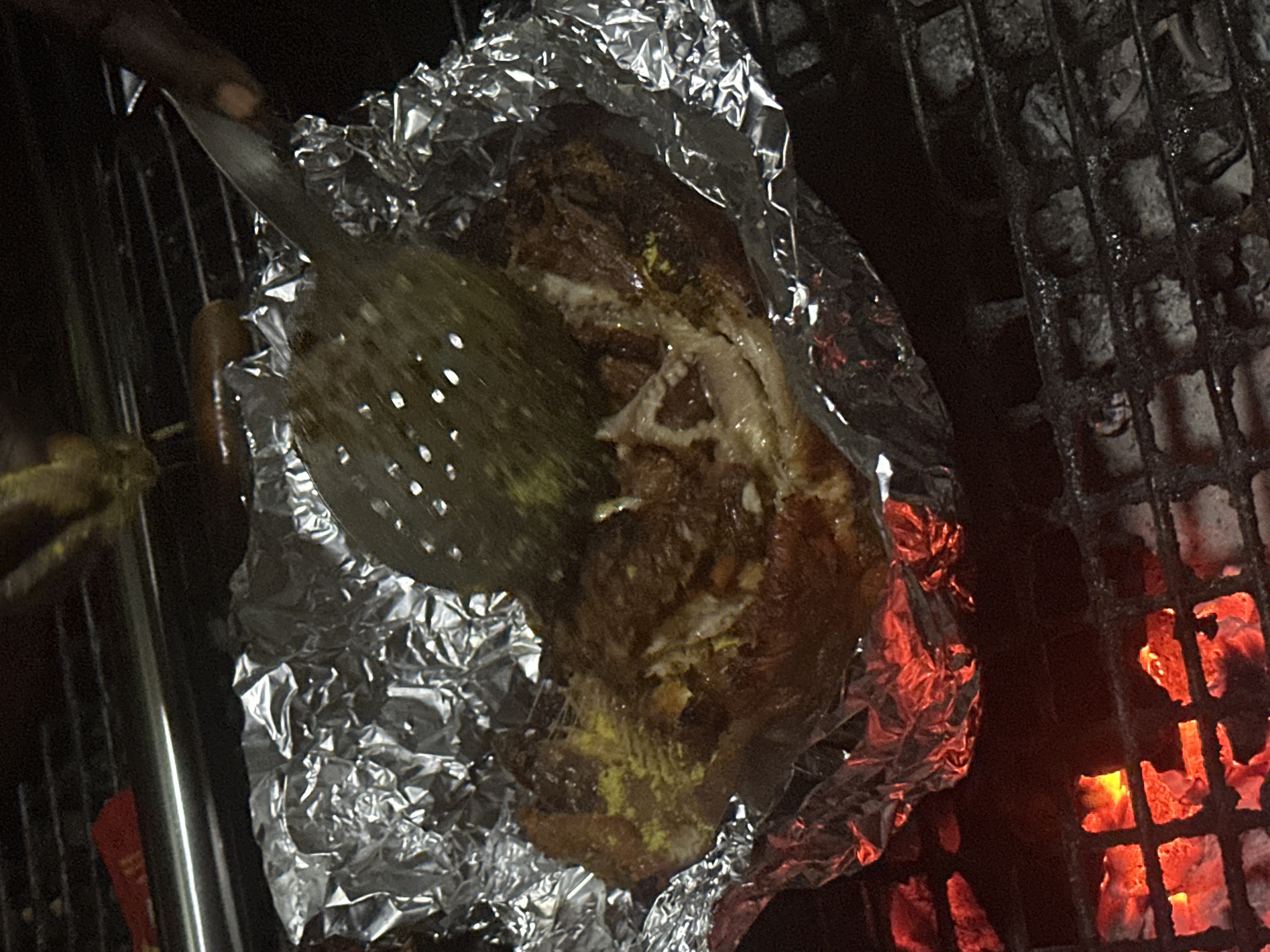 A piece of food, possibly chicken or pork, is being cooked on a grill. It is wrapped in aluminum foil and appears to be seasoned with a yellow spice. A slotted spatula is positioned above the food, as if to turn or check it. The food is placed on a grill rack, with glowing red embers visible below, indicating it is over a charcoal fire. The scene suggests an outdoor cooking setup, possibly in Jos, Nigeria, given the context provided, though the specific location within the city cannot be determined from the image alone. The overall impression is of a rustic and informal grilling experience.