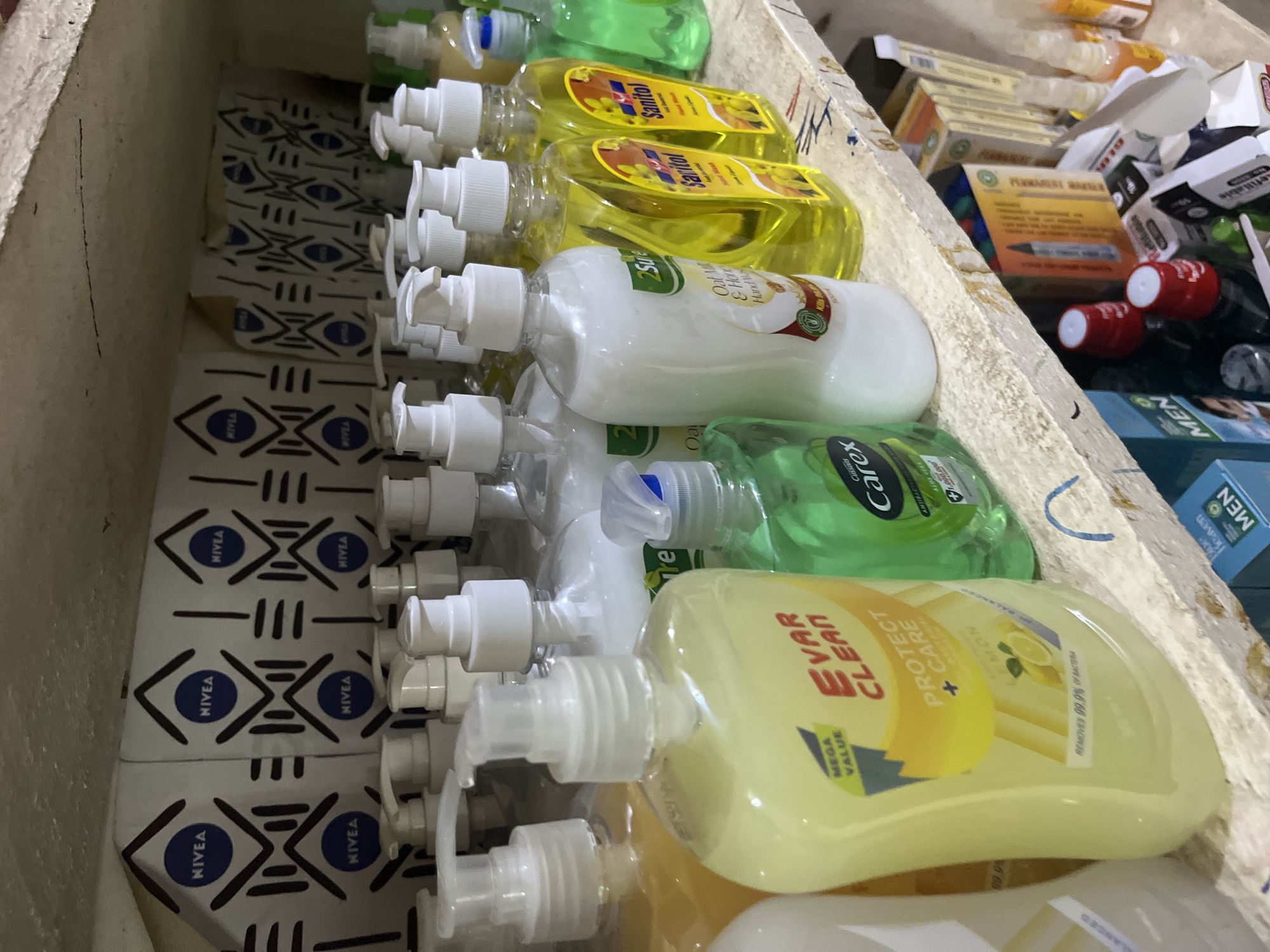 The image shows a shelf stocked with various liquid soap and hand sanitizer bottles. In the foreground, a yellow bottle of "Evar Clean Protect + Care" hand wash with lemon scent is prominently displayed. Behind it, several other bottles of liquid soap and hand sanitizer are visible, including brands like "Sanitol" and "Carex". The bottles have white pump dispensers and clear plastic containers, with labels indicating different scents and functions. The shelf itself is made of a light-colored material, possibly wood or particle board, and appears somewhat worn. The background features a patterned cloth with repeated "NIVEA" logos. The overall impression is that of a retail display, likely in a small shop or market stall in Bukuru, Nigeria, given the context. There are no people visible in the image.