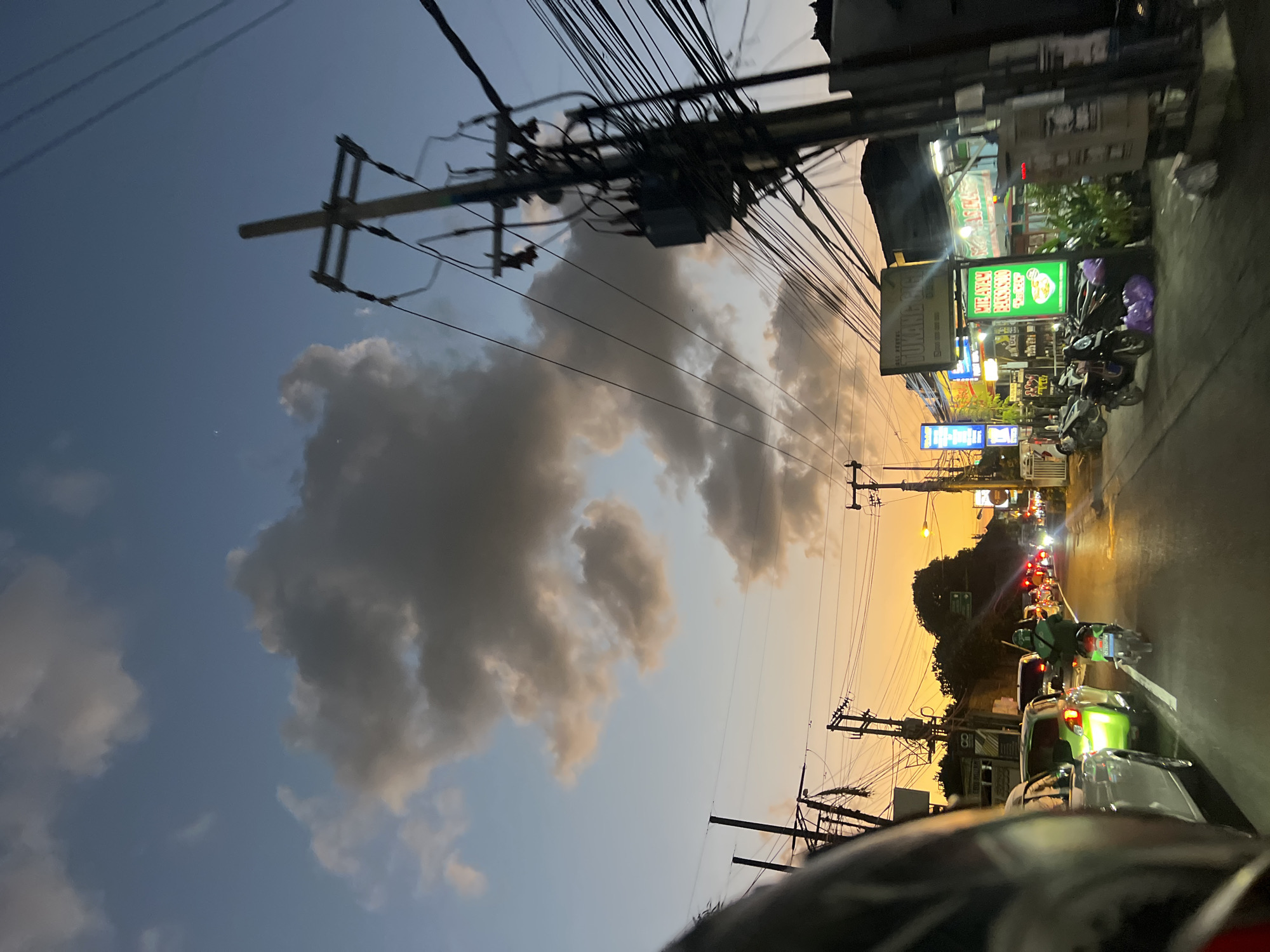 The image captures a street scene in Denpasar, Indonesia, during twilight. The sky is a gradient of deep blue, transitioning to a soft orange hue near the horizon, with scattered clouds tinged by the setting sun. Power lines crisscross the frame, supported by utility poles, creating a sense of urban complexity. A stream of traffic moves along the road, including cars and motorcycles. One motorcycle rider, wearing a green jacket and helmet, is visible in the foreground. Shops and businesses line the street, some illuminated by neon signs, one of which displays text in Indonesian, including "AYAM BETUTU CHICKEN." Several motorcycles are parked on the sidewalk. The overall impression is a typical evening in a bustling Indonesian city.