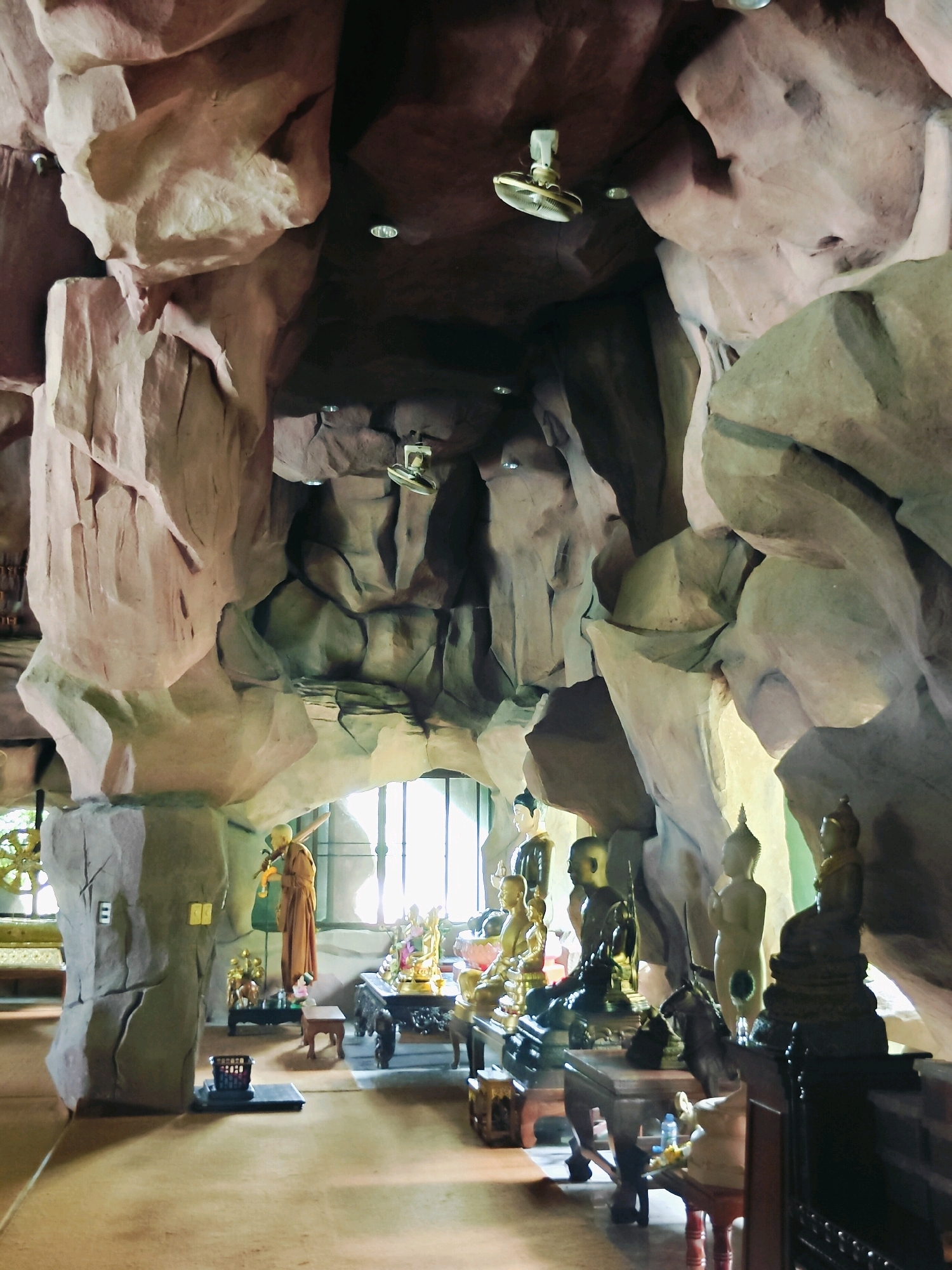 An interior shot shows the cavernous space of a Buddhist temple. The walls and ceiling appear to be made of sculpted rock formations, with some areas resembling smooth, worn stone and others exhibiting a rough, textured surface. Several ceiling fans are visible, mounted against the dark brown ceiling. The floor is a smooth, light brown surface.
In the foreground, a collection of Buddha statues of varying sizes and materials, including gold and dark bronze, are arranged on tables and platforms. Some statues are standing, while others are seated. A monk in an orange robe stands near a window, holding a long, thin object. Behind him, through a large window with vertical bars, daylight illuminates the scene. Various decorative elements, such as small tables, baskets, and what appear to be offerings, are scattered throughout the space. The overall impression is one of a sacred and somewhat rustic environment, possibly a cave adapted for religious worship.