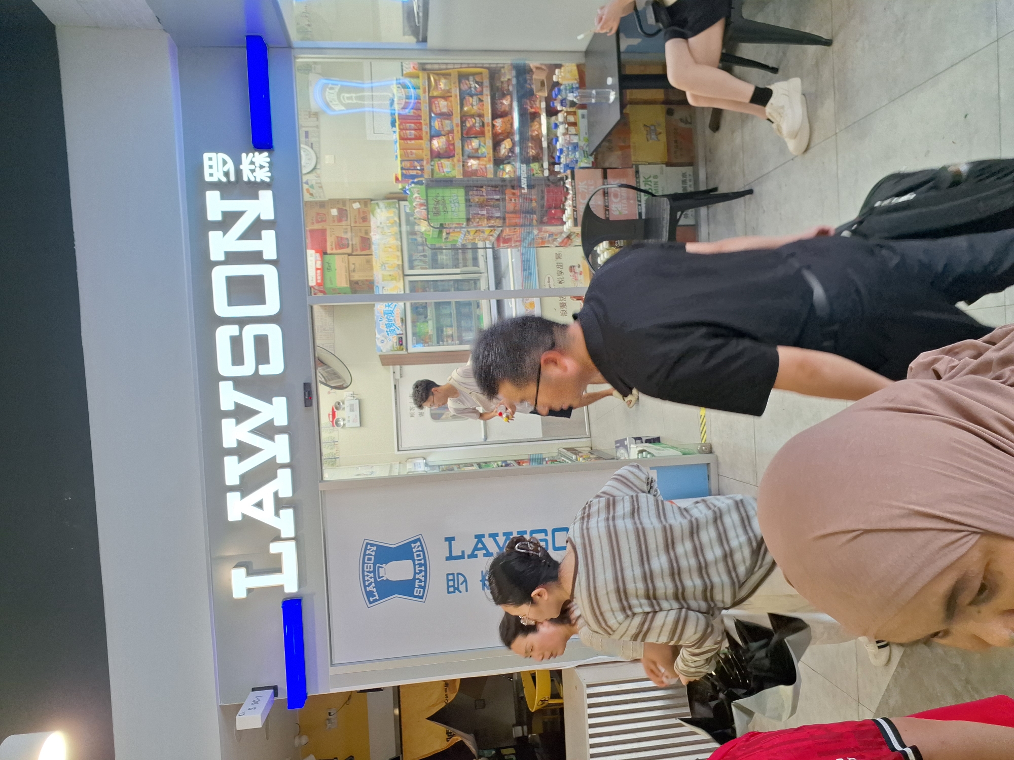 The image depicts the entrance of a Lawson convenience store in Hongkou District, China. A bright, illuminated sign with the word "LAWSON" in large white letters is prominently displayed above the entrance. To the right of "LAWSON", Chinese characters are visible. Below the main sign, a smaller Lawson Station logo is visible on the glass door. Inside the store, shelves are stocked with various products, including drinks and snacks. Several people are visible in the scene. In the foreground, a person wearing a black t-shirt and pants is walking away from the camera, carrying a black bag. To the left, two women are standing, one wearing a striped long-sleeved shirt and the other in a tan headscarf. Another person, a man in a white t-shirt, is standing inside the store near the shelves. In the background, a person is seated at a table with a black chair next to them. The floor appears to be tiled.