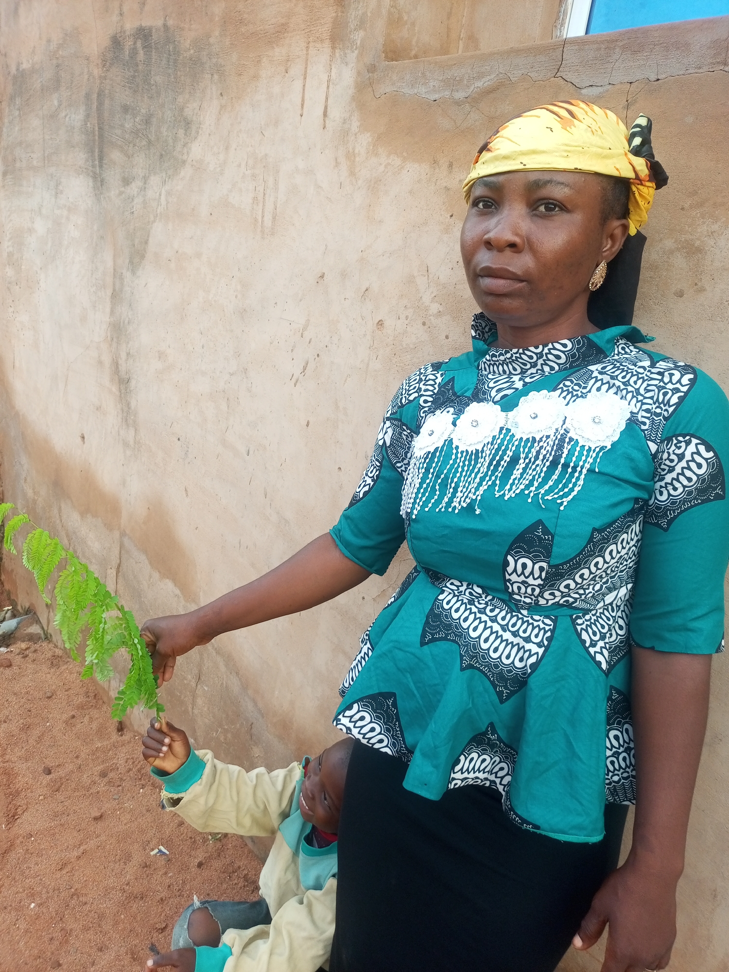 In Tempure, Nigeria, a woman stands leaning against a textured, light brown wall. She wears a yellow headscarf with black and brown patterns and a teal peplum top adorned with white floral embellishments and black and white geometric prints. She is holding a sprig of green leaves in her right hand, reaching towards a child who is partially visible in the lower left corner of the frame. The child is wearing a light tan jacket with teal accents and appears to be looking up at the woman. The ground in front of them is covered in reddish-brown dirt. The wall behind them has a rough, uneven surface with some darker discolorations.