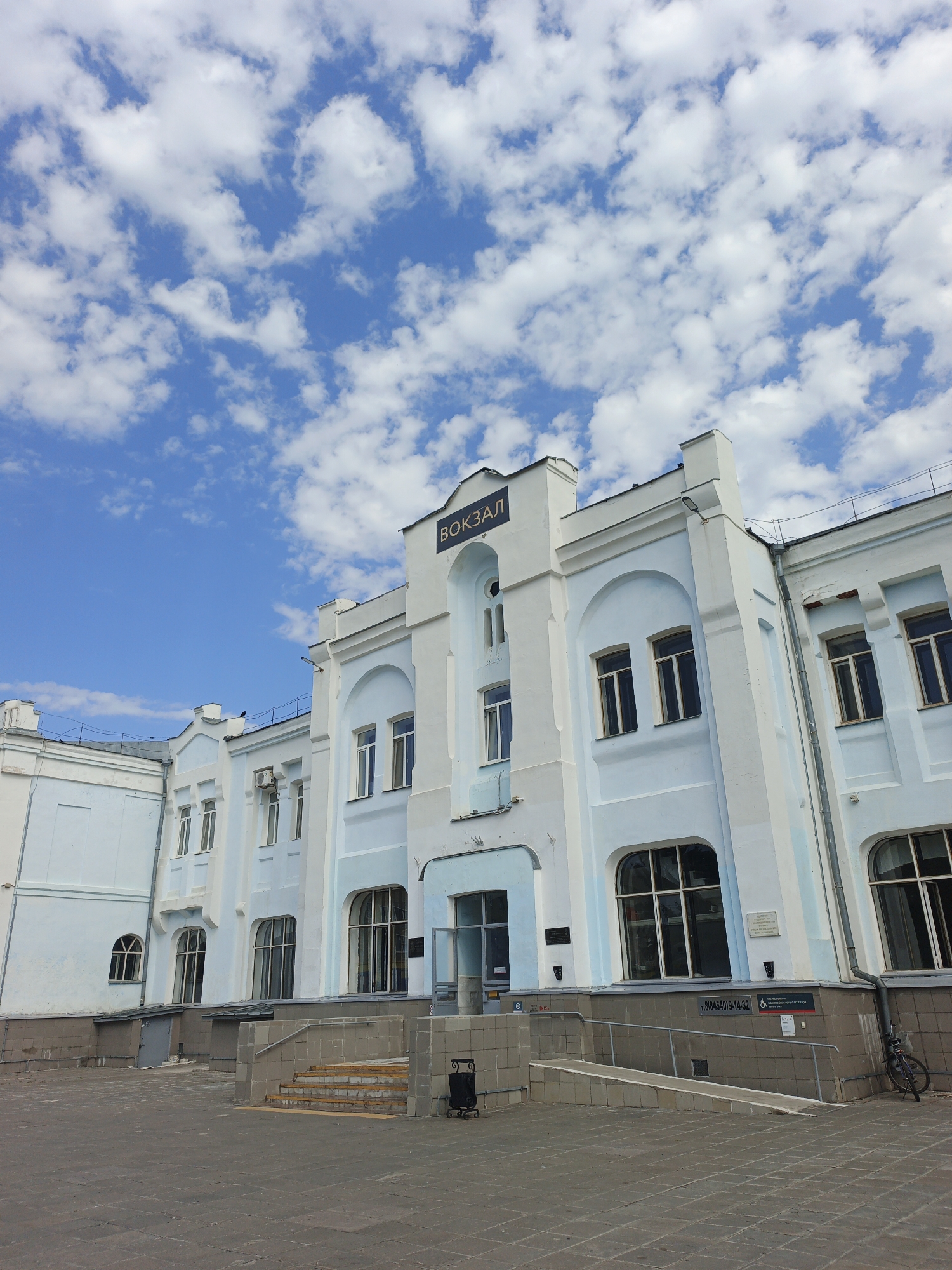 The image depicts the exterior of a light blue and white building, identified by the Cyrillic word "ВОКЗАЛ" (Vokzal) meaning "train station" on its facade. The building has multiple windows, some arched and some rectangular, with dark frames. The architecture appears somewhat dated, with decorative elements and cornices. In front of the building, there is a paved plaza. A set of stairs and a ramp lead up to the entrance. To the right of the entrance, a bicycle is parked against the building. The sky above is blue with scattered white clouds. The overall scene is one of an operational train station on a clear day. Based on the signage and architectural style, this is likely the train station in Rtishchevo, Russia.