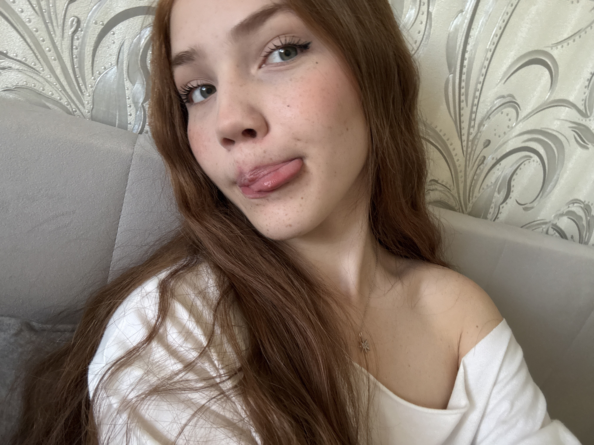 A close-up selfie captures a young woman with long, wavy reddish-brown hair, fair skin dotted with noticeable freckles, and light-colored eyes, appearing green or hazel. She is seen from the chest up, wearing a light-colored, possibly off-white, top that partially exposes her right shoulder. A delicate silver-toned necklace with a small, star-shaped pendant is visible around her neck.
The woman is looking directly at the camera with a playful and mischievous expression, sticking her tongue out slightly. Her long, dark eyelashes are prominent, contrasting with her light eye color.
The background reveals an indoor setting. To her left, a section of a light gray upholstered sofa or headboard is visible. To her right, the wall is covered with a decorative wallpaper featuring an elegant, metallic silver or gray floral and scroll pattern on a lighter, possibly white or cream-colored, background. The lighting is soft and even, suggesting a daytime shot. No specific time of day or weather conditions are discernible beyond the indoor context. The image conveys a casual and cheerful mood. There is no visible text. This scene is located in Naberezhnye Chelny, Russia.