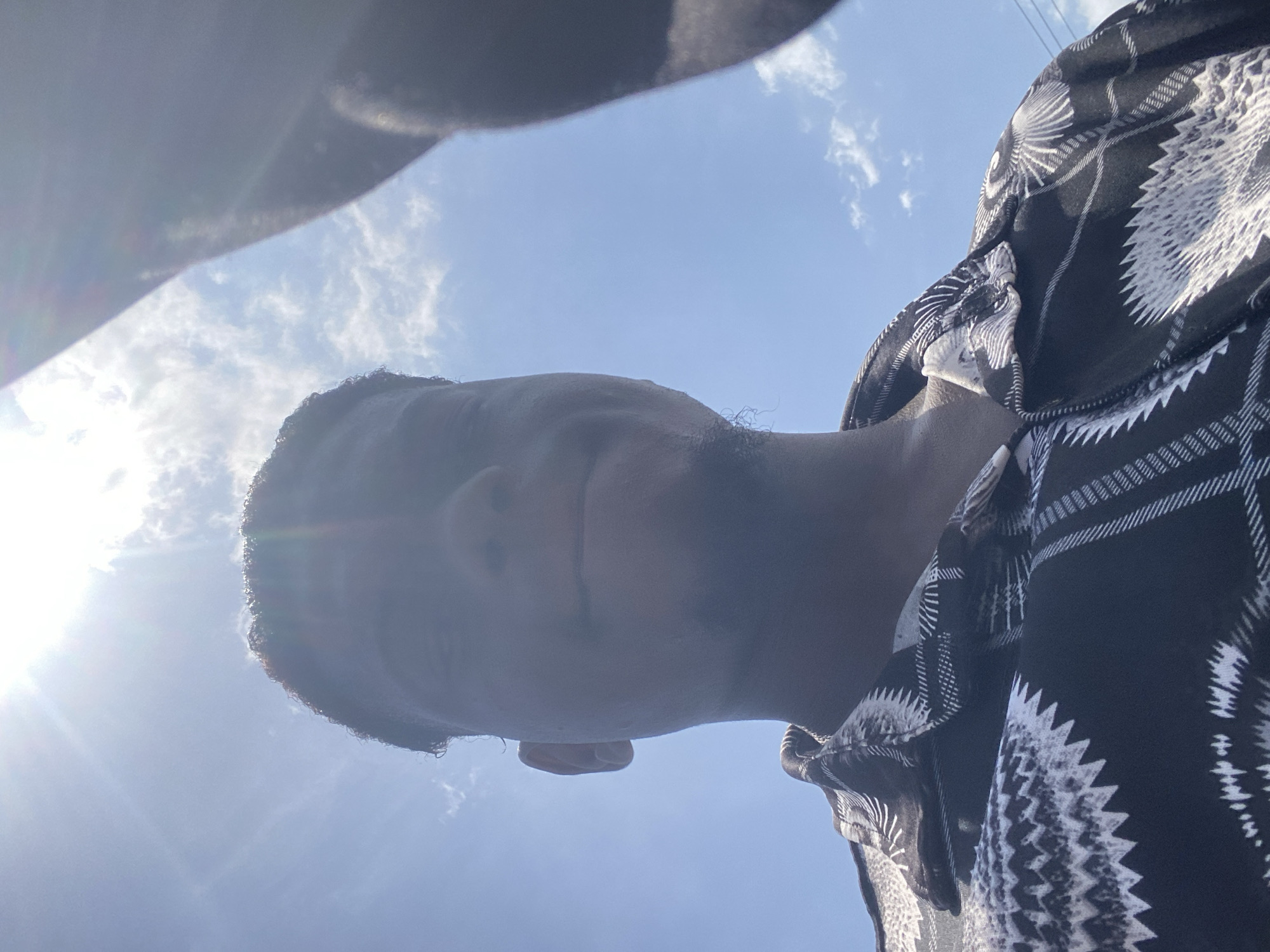 An upward-angled portrait captures a person, likely male, from the chin upwards, silhouetted against a bright blue sky with scattered white clouds. The subject's head is slightly tilted to the right, and their face is largely in shadow, with a neutral expression. A strong sun flare emanates from the upper left corner, indicating bright daylight. They are wearing a dark shirt, possibly black, with a distinctive white pattern that appears geometric or abstract, featuring sharp, spiky, and wavy designs. The perspective is from below, looking up, giving a sense of the sky stretching behind and around the person. No specific location cues are visible beyond the sky and clouds. There is no visible text.
