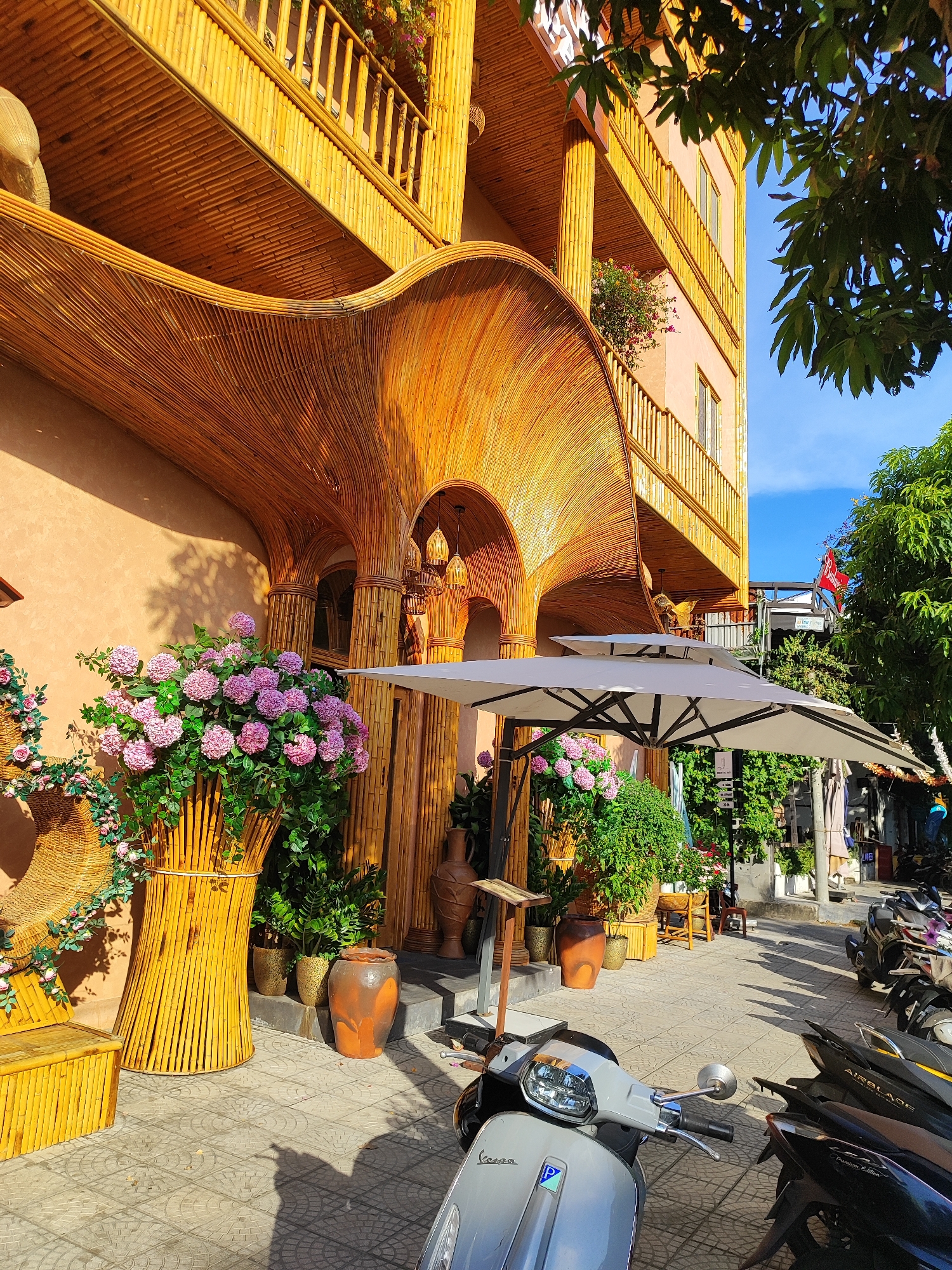 A light gray Vespa scooter is parked in the foreground on a tiled sidewalk. Behind the scooter, a row of dark-colored scooters is parked along the street. The street is lined with buildings and lush green trees.
To the left of the sidewalk is a building with a facade constructed from bamboo. The bamboo is used for both structural elements, like columns and arches, and decorative purposes, such as the curved entryway canopy. Large potted plants with pink flowers are placed in front of the bamboo structure. A wooden bench is also visible to the left.
The building appears to be a multi-story establishment with balconies adorned with bamboo railings. The overall architectural style suggests a tropical or Southeast Asian influence. The lighting indicates it is daytime, with sunlight casting shadows on the ground. The scene is consistent with a commercial area in a city, likely a cafe or shop, in Da Nang, Vietnam.