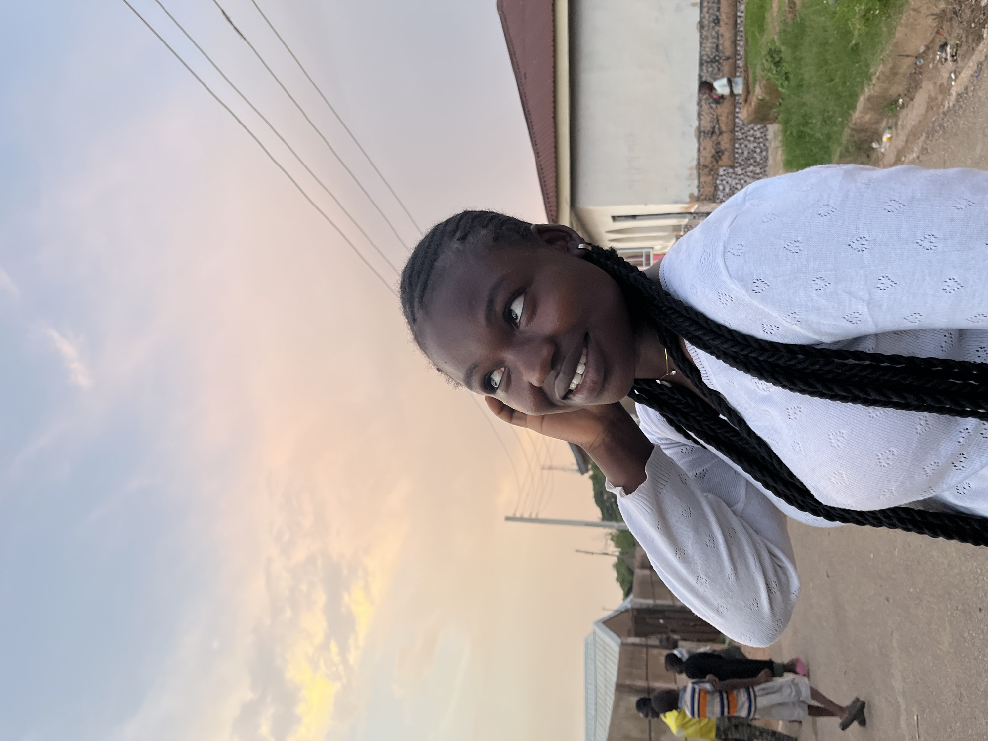 An outdoor, eye-level, medium shot features a young Black woman looking upwards and to the right, with her right hand touching her ear. She has long, black braids that fall over her shoulders and a white, long-sleeved shirt with a textured pattern. Her left arm is bent and resting at her side.
In the background, a dusty, unpaved road is visible, with several people in the distance. To the left, there are small, concrete buildings with corrugated metal roofs. To the right, a larger building with a brown, corrugated metal roof and white walls is present. The sky is a soft blend of pale blue and pastel orange, suggesting either sunrise or sunset. Power lines are visible crisscrossing the upper right portion of the frame. The overall setting appears to be a residential area in Jos, Nigeria.