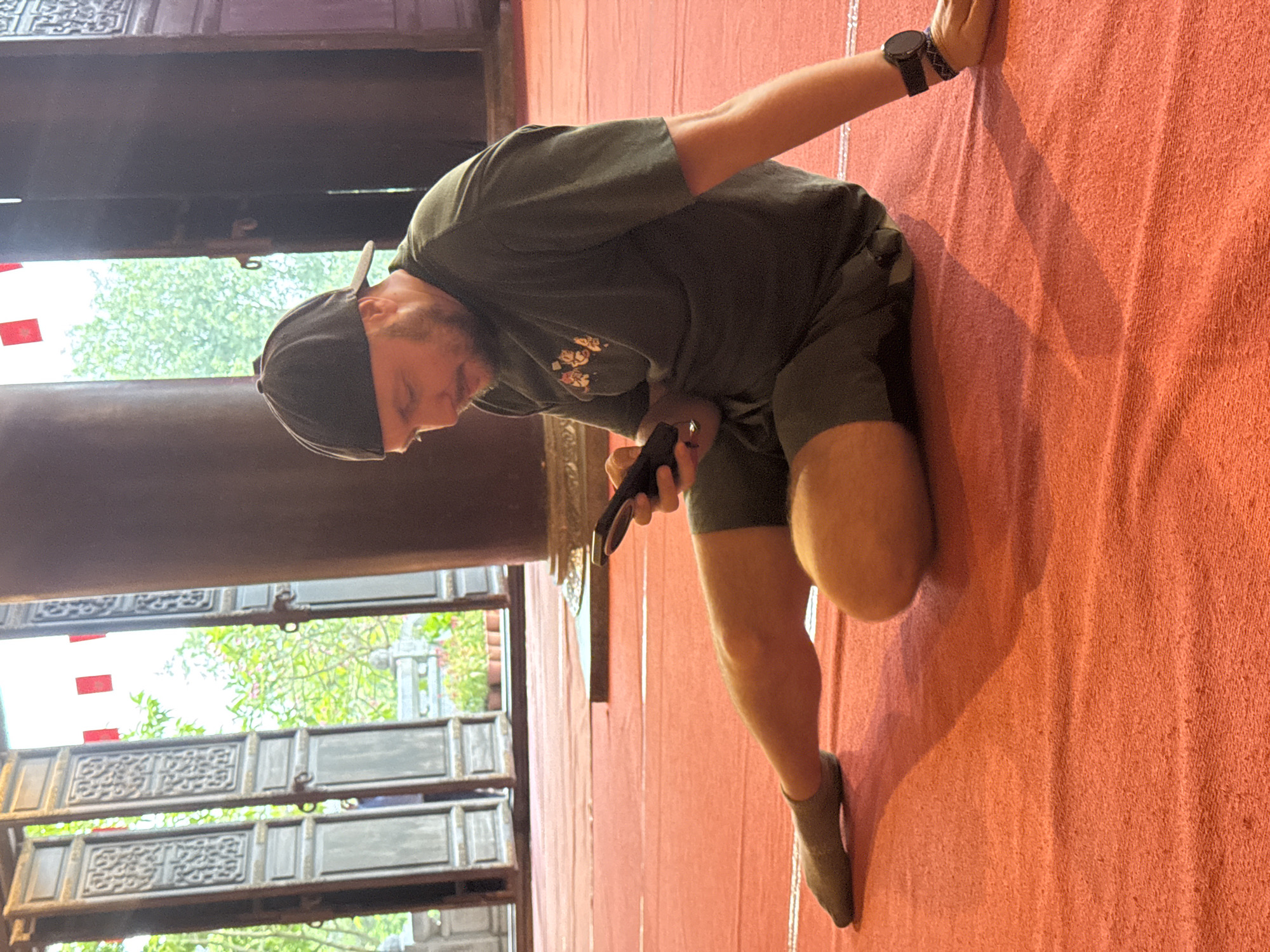 A man wearing a dark green t-shirt, dark shorts, a black baseball cap, and grey socks is sitting on a red carpeted floor, looking at his phone. He is indoors, with large wooden doors and columns visible in the background. The doors are open, revealing greenery and what appears to be a bright sky. Red fabric is hanging from the ceiling. The man is positioned on the right side of the frame, with his legs extended. A smartwatch is visible on his left wrist. The setting suggests a temple or a traditional building in Tây Hoa Lư Ward, Vietnam, given the architectural style and the presence of potted plants.