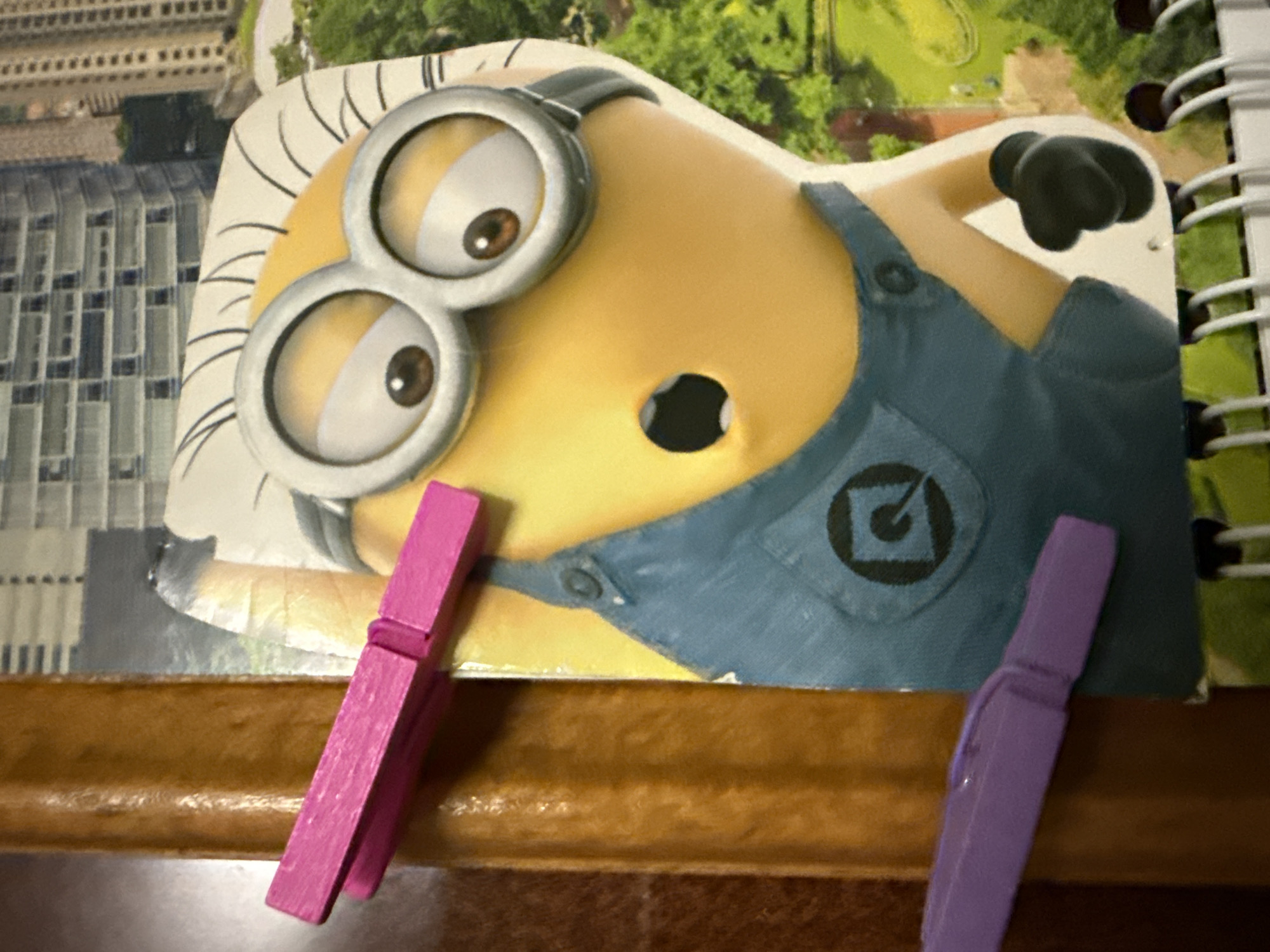 A close-up shot shows a Minion character cutout attached to a spiral-bound notebook. The Minion is depicted wearing blue overalls and its signature goggles. A pink clothespin is clipped to the notebook's spine, positioned just above the Minion's head. Below the Minion, a purple clothespin is also clipped to the spine. The background is blurred, but appears to be a cityscape with buildings and trees. The lighting is bright and even, suggesting an indoor setting.