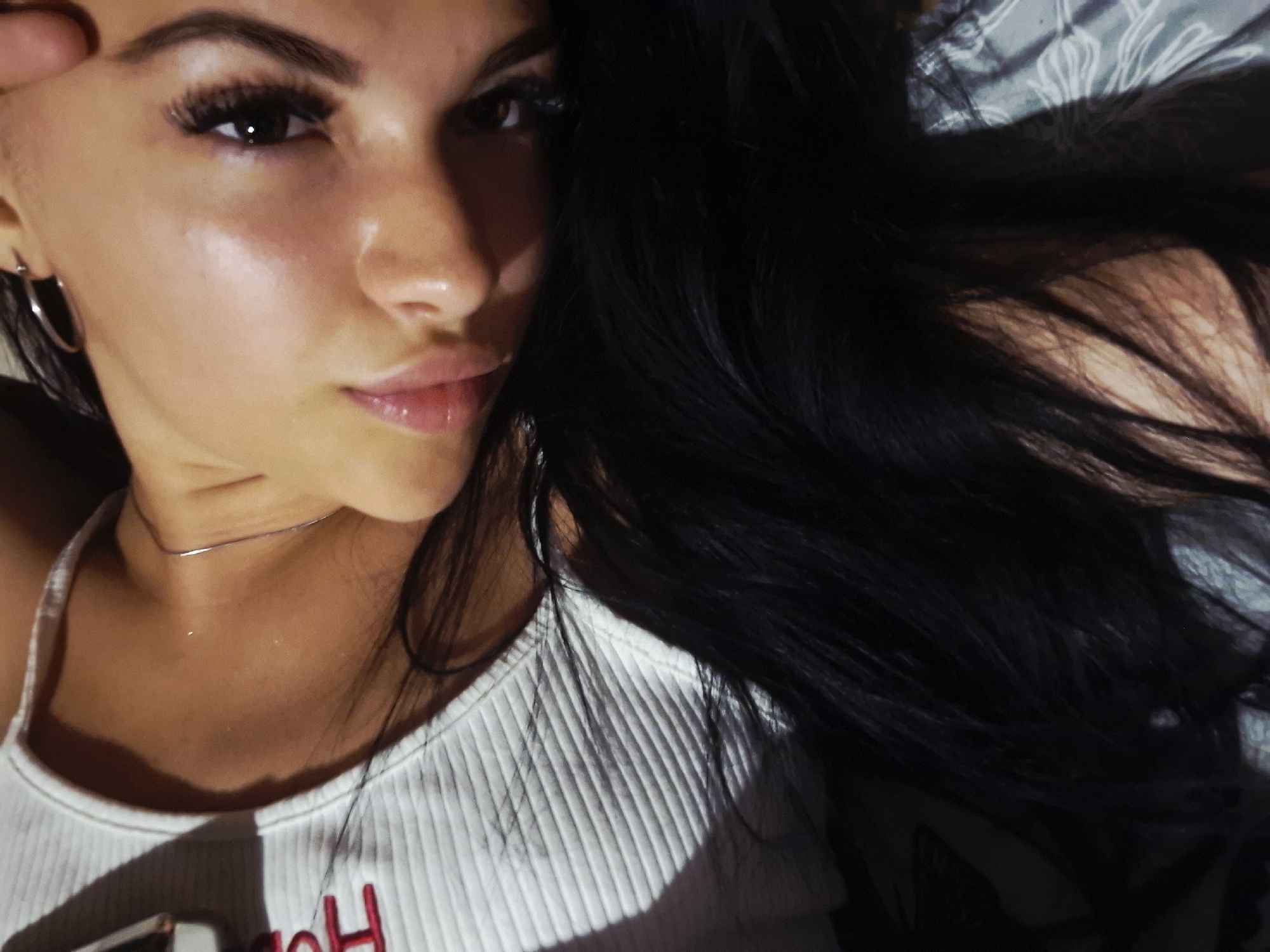 A close-up portrait shows a young woman lying down in an indoor setting, likely a bedroom, in Krapets, Bulgaria. She has dark, long hair, which cascades around her head and covers part of the frame. Her face is turned slightly towards the viewer, with her dark eyes looking directly into the camera. She has full, glossy lips, well-defined eyebrows, and prominent dark eyelashes. A light source from the left illuminates her face, highlighting her cheek and nose, and creating soft shadows.
She wears a simple, thin silver choker necklace and a silver hoop earring in her visible left ear. Her attire consists of a white, ribbed tank top or camisole. On the lower left part of her top, a red, stylized letter, possibly an "H", is partially visible. The background features a patterned fabric, possibly a bedsheet or pillowcase, in grey and white tones. The overall lighting suggests an evening or night-time environment. Her expression is calm and direct.