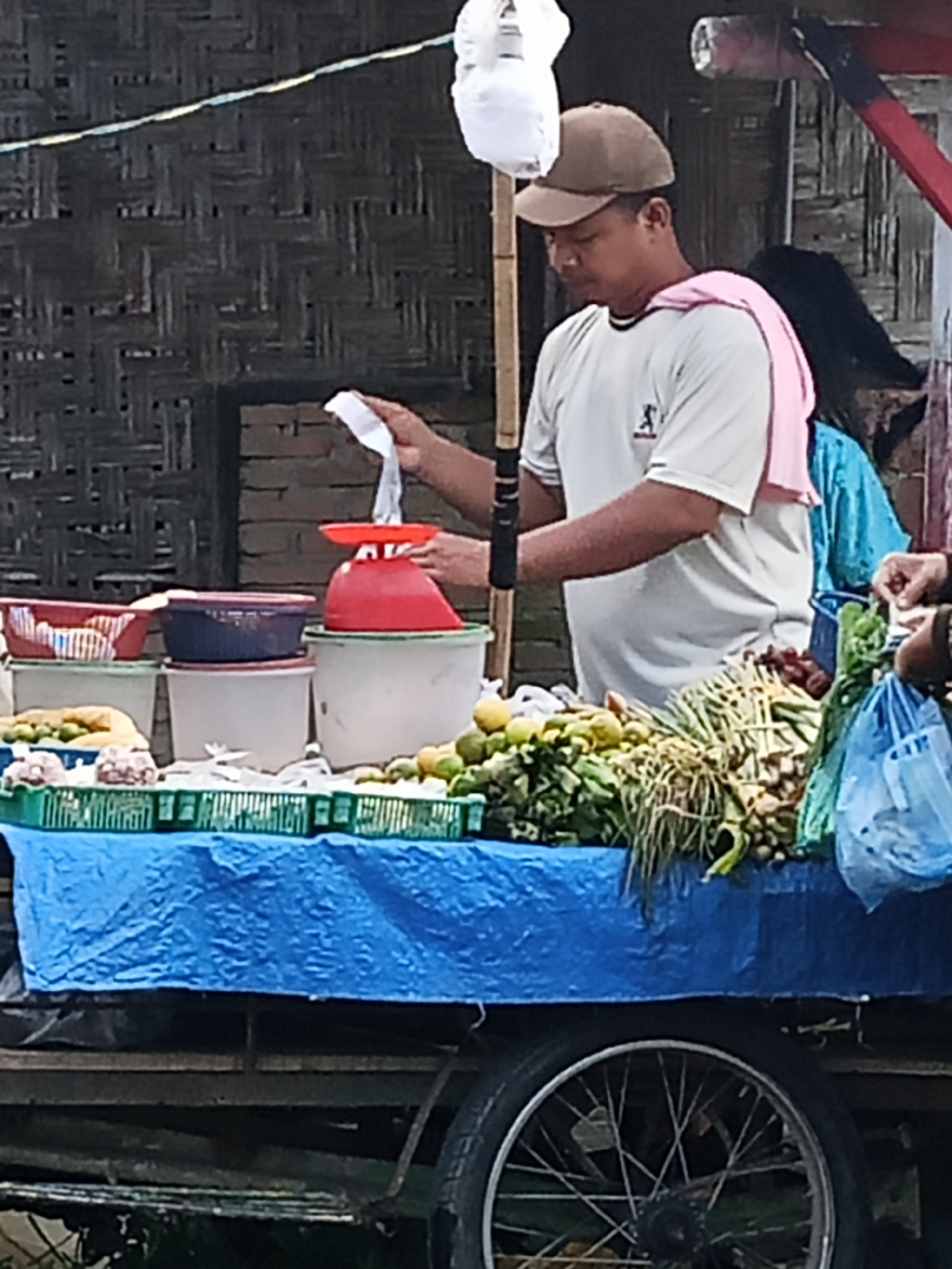 In Wono Sari, Indonesia, a man wearing a light-colored t-shirt and a brown cap is standing behind a cart filled with produce. He is holding a white piece of paper and appears to be in the process of weighing items on a red scale. The cart is covered with a blue tarp, and various containers and baskets of fruits and vegetables are visible. Behind the man, a person wearing a blue garment is partially visible. The background consists of a woven bamboo wall. The scene depicts a street-side market stall.