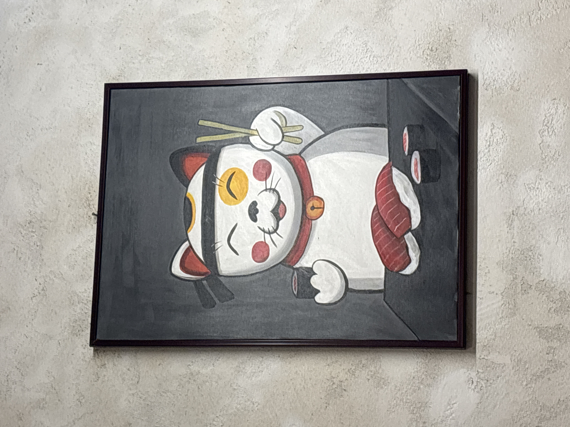 The image shows a painting of a cartoon cat holding chopsticks and sushi, framed and hanging on a textured wall. The cat is white with red and yellow accents on its head and face, and it is wearing a red collar with a bell. It is holding a piece of sushi in its left paw and chopsticks in its right paw. In front of the cat, there is a plate of sushi and two rolls of sushi. The background of the painting is dark gray. The wall appears to be a light, neutral color with a rough texture. The painting is hanging from a small nail on the wall. No specific location in Đà Nẵng, Vietnam is identifiable from the image.