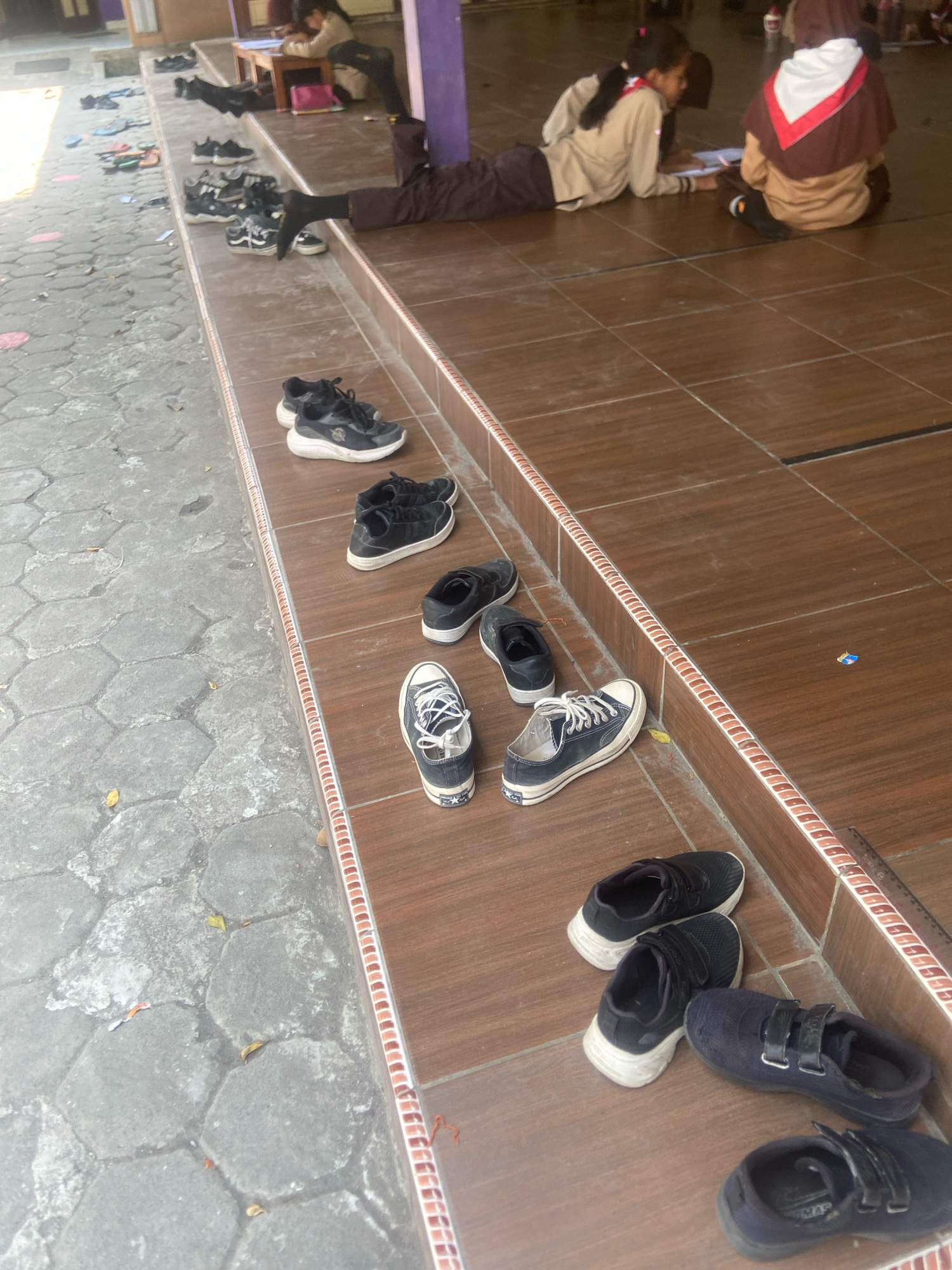 The image depicts an outdoor scene on a tiled surface adjacent to a paved area, likely in Sleman, Indonesia. The tiled area is brown with a decorative border. In the foreground, on the tiled surface, several pairs of shoes are arranged. These include black sneakers with white soles, black low-top canvas shoes with white laces, and dark blue velcro shoes. Behind the shoes, two young girls are visible. One girl, wearing a tan uniform with a red and white neckerchief, is lying on her stomach and appears to be writing or drawing on a piece of paper. Another girl, also in uniform with a tan top and a brown hooded garment, is seated beside her, facing away from the camera. The paved area to the left shows more shoes scattered, some appearing to be school footwear. In the background, there are glimpses of a building and additional people. The overall impression is of a school or educational setting where students are engaged in an activity outdoors.