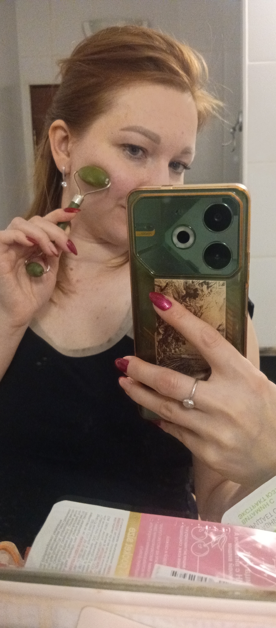 A woman with fair skin and auburn hair is taking a selfie in what appears to be a bathroom. She is holding a green jade roller to her face with her left hand, and her right hand is holding a smartphone, which has a green case and a botanical image on the back. She wears a black tank top and a ring on her right ring finger, and her nails are painted a dark red color. In the foreground, there is a package of a cosmetic product with some text in another language. The environment includes a mirror and possibly a tiled wall in the background. The city and specific location are not discernible.