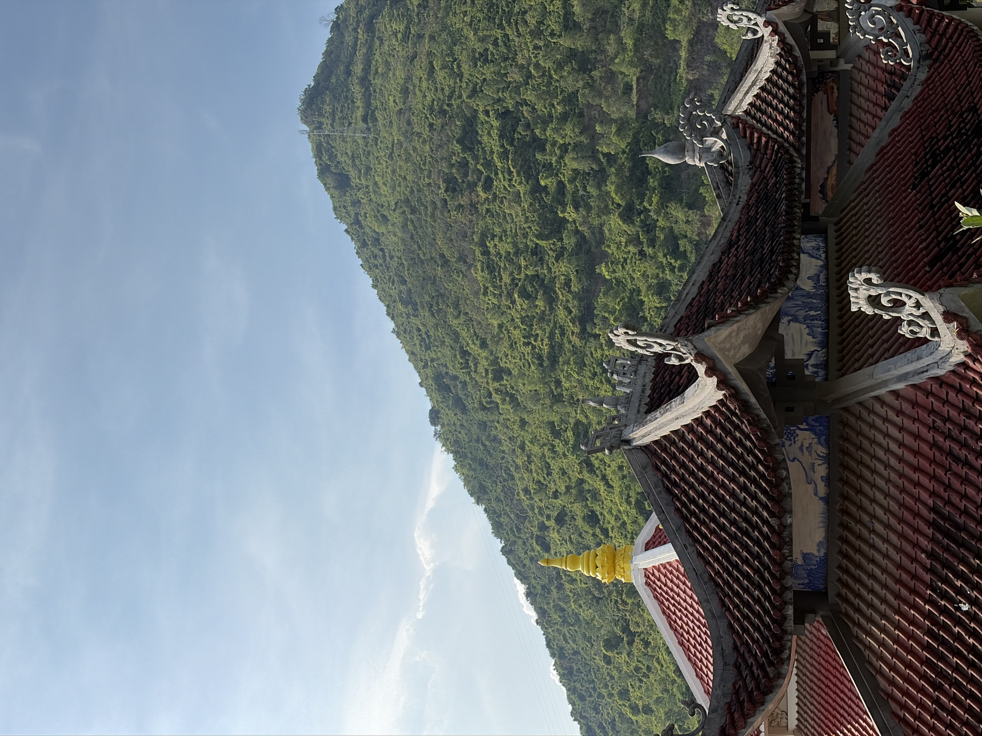 The image shows a close-up view of traditional Vietnamese temple rooftops in Tay Nha Trang Ward, Vietnam, with a lush, green mountainside in the background under a clear blue sky with some wispy clouds. The foreground is dominated by the intricately designed, reddish-brown tiled roofs. These roofs feature ornate white and gray decorative elements along the edges, resembling stylized waves or clouds. One of the roofs has a pointed, bright yellow spire that stands out against the darker tiles. Below the ornate edges, there are sections with blue and white painted murals depicting landscapes. The mountain behind the temple is densely covered in green vegetation, with a visible transmission tower near its summit. The overall scene conveys a sense of cultural heritage and natural beauty.