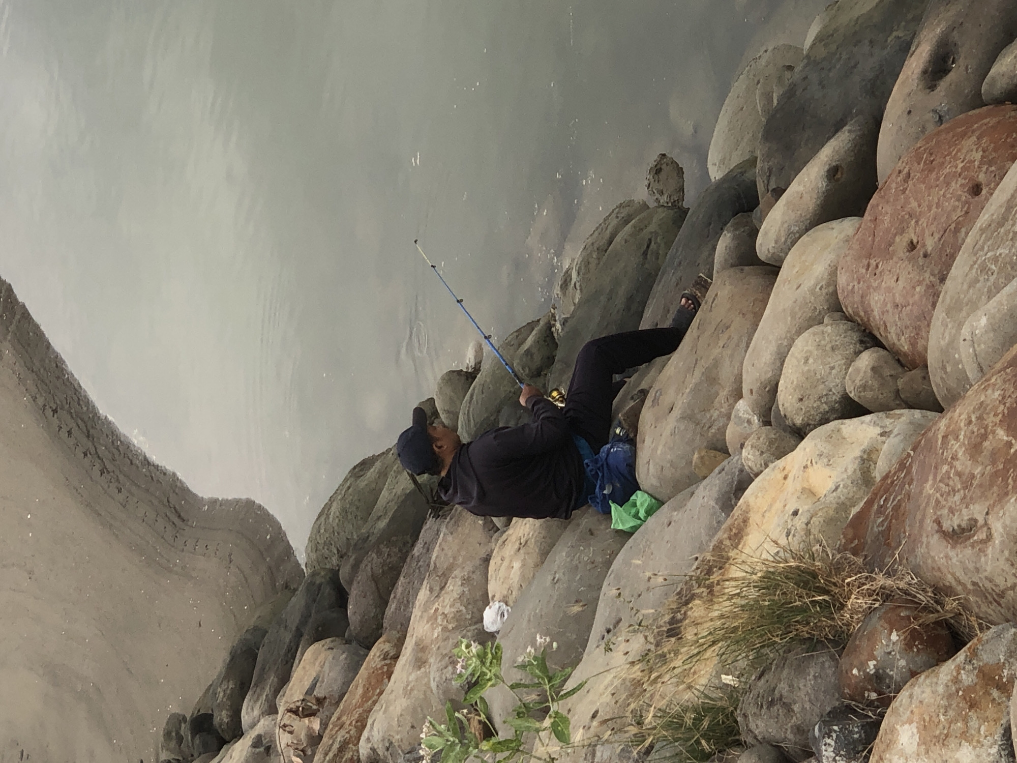 An individual is seated on a rocky shoreline, facing away from the camera and towards a body of water, engaged in fishing. The person is wearing a dark hooded jacket, dark pants, a baseball cap, and sandals. A blue fishing rod is held in their hands, with the line cast into the water. Next to the individual, on the rocks, are a blue and green bag. The shoreline is composed of large, varied-colored rocks, with some sparse vegetation growing among them, and a sandy bank to the left. The water appears calm and reflects the overcast sky. This scene likely depicts the recreational area along the coast of Lhokseumawe, Indonesia.