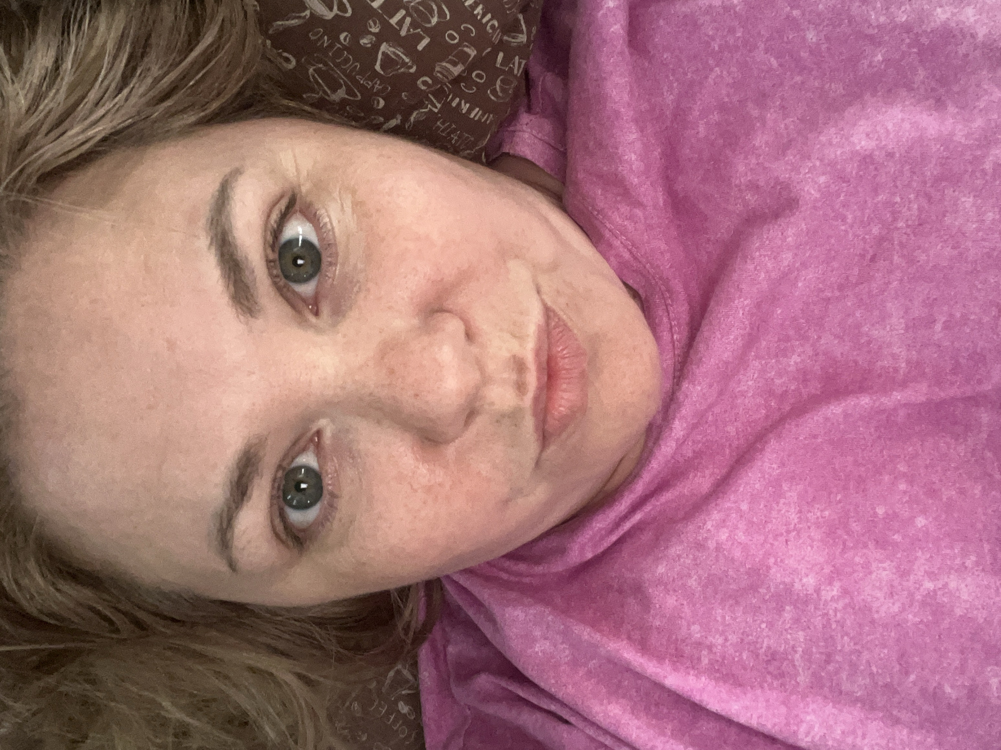 A close-up, angled shot of a woman's face. She has fair skin, green eyes, and blonde hair. She appears to be wearing a pink shirt or top. The background includes a pillow with a coffee-themed pattern. The image does not provide information to determine a specific location in Vesyoloye, Russia, or to identify actions, interactions, or details about the environment beyond what is immediately visible.