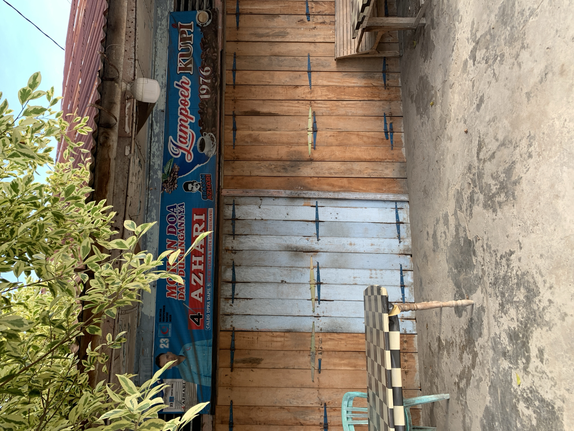 The image captures the exterior of a small establishment in Meunasahlhok, Indonesia, with a focus on a wooden facade and a banner above. The building appears to be a shop or restaurant, indicated by the banner that reads "MOHON DOA DAN DUKUNGANNYA 4 AZHARI Lampoch Kupi 1976". This suggests it is a coffee shop named "Lampoch Kupi" established in 1976, and likely a campaign banner for a candidate named Azhari.
The facade consists of vertical wooden planks, with a section painted a light blue on the right. Blue metal hinges are visible along the seams of the wooden doors. In front of the building, there is a checkered table with a black and white pattern, supported by a carved wooden leg. A teal chair is placed on the left side of the table. A wooden bench with a similar checkered pattern is situated on the right side of the image, partially obscured by the table. The ground is a concrete surface, showing signs of wear and some debris. Overhead, the branches of a leafy tree with variegated green and white leaves cast shadows on the scene, suggesting it is daytime with dappled sunlight. The sky is visible in the upper portion of the image, appearing clear and blue.
