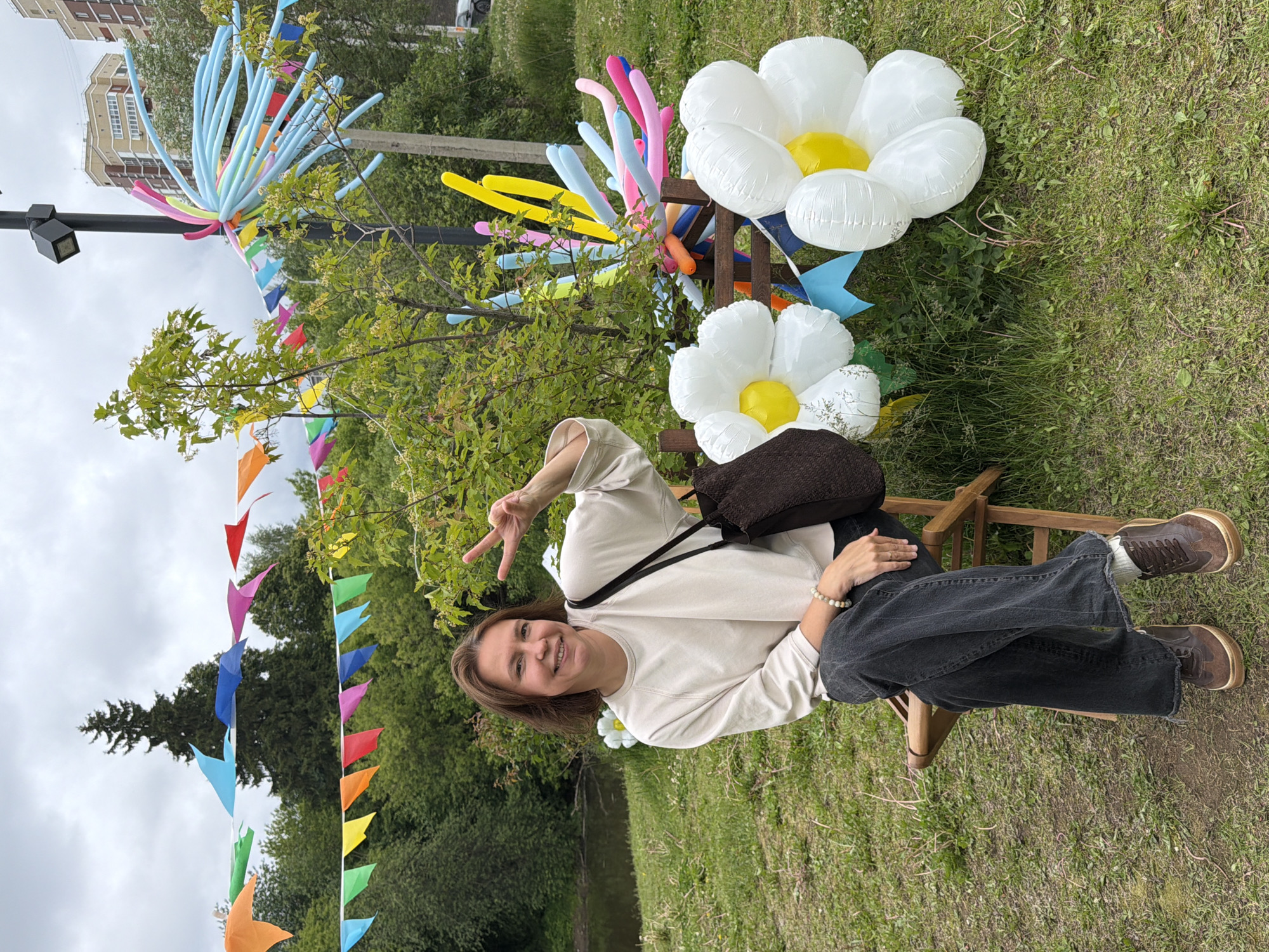 An outdoor scene in the city of Izhevsk, Russia, depicts a woman seated on a wooden folding chair amidst festive decorations. The woman, with short brown hair, is wearing a light-colored long-sleeved top, dark wide-leg jeans, and brown sneakers. She smiles at the camera, holding up her right hand in a peace sign. A dark brown tote bag hangs from her left shoulder, and a light-colored beaded bracelet is on her left wrist.
The ground is covered with green grass. To the left and behind the woman, a body of water is visible. Lush green trees and bushes form a dense background across the scene. Various colorful triangular pennant flags are strung horizontally across the upper portion of the image, between the trees and a black lamppost.
Near the lamppost on the right, a large decorative arrangement of long, thin balloons in shades of blue, pink, and yellow emanates outward, resembling an exploding shape. On the grass to the right of the woman, two large white inflatable flowers with yellow centers are present, with a third partially visible behind her to the left. Behind these flowers, another balloon arrangement with similar long, colorful balloons is visible, supported by a wooden structure. In the distant background, multi-story residential buildings are visible under an overcast sky, with a dark vehicle partially visible on a road.