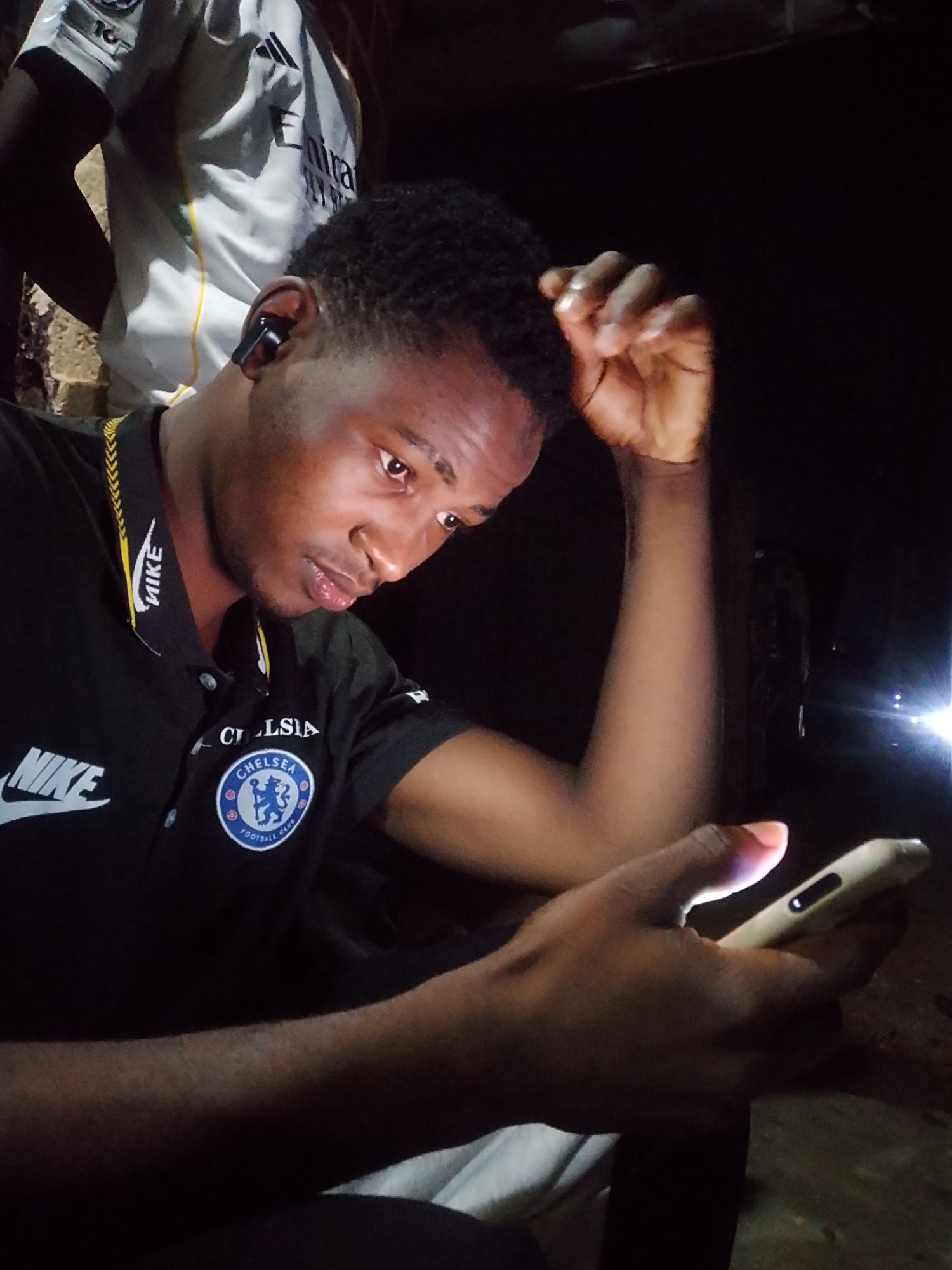 A medium shot, taken at night in an outdoor urban setting, captures a young Black male seated, looking downwards at his smartphone. He is wearing a black polo shirt with a Chelsea Football Club logo and a Nike emblem. He has short, curly dark hair and is wearing an earbud. His right arm is bent, with his hand resting on his head. The background is dark, with a hint of a brick wall and another person in a white shirt visible. The lighting is dim, with the phone screen providing illumination.