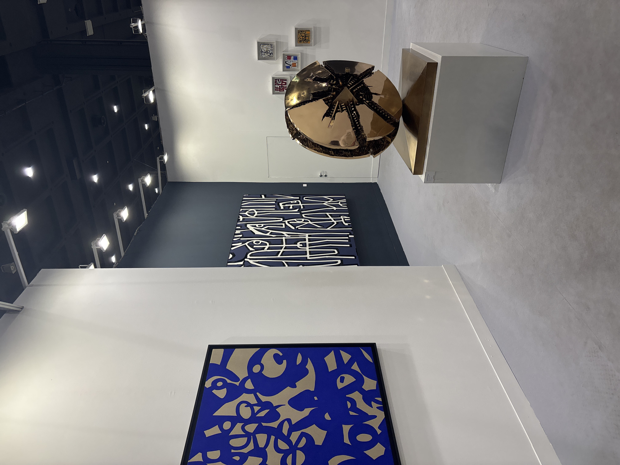 This image captures an interior view of an art gallery or exhibition space, likely in Milan, Italy, showcasing various modern artworks under artificial lighting.
In the mid-ground, a striking, large, fragmented golden sphere sculpture dominates the scene. The sphere appears to be made of polished metallic segments that are deliberately broken or pulled apart, revealing an intricate, dark, complex interior structure. It rests on a simple white rectangular pedestal. Above the sphere, on a white wall, are four smaller, framed artworks, each featuring abstract or calligraphic designs within white frames.
Further left, on a dark grey accent wall, hangs a large, rectangular painting composed of thick white, angular, and curvilinear lines against a dark blue background, creating an abstract pattern. In the immediate foreground, another large, rectangular artwork with a black frame displays bold, flowing blue abstract shapes and organic lines set against a light beige or tan background.
The gallery space features clean white walls, a dark grey accent wall, and a high, dark ceiling fitted with numerous square recessed spotlights, illuminating the artworks. The floor is light-colored and minimalist. A subtle reflection of a person can be observed on the polished surface of the golden sphere, suggesting the presence of a viewer or the photographer. The overall atmosphere is one of modern art display and appreciation. No discernible text is visible on any of the artworks or surrounding signage.