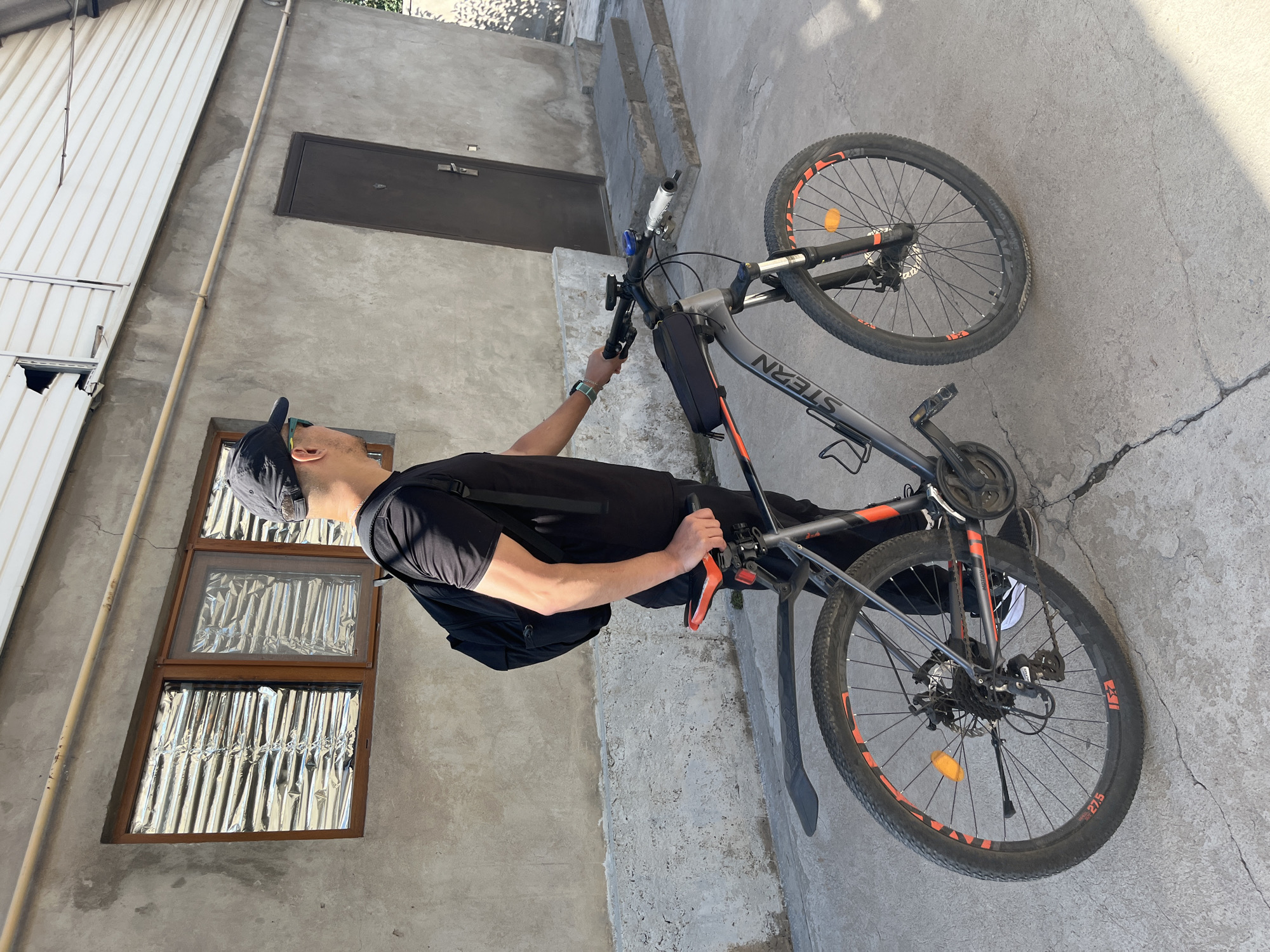 In Shymkent, Kazakhstan, a man wearing a black t-shirt, black pants, white sneakers, and a black baseball cap stands next to a bicycle. He has a black backpack on his shoulders and is holding the handlebars of the bicycle. The bicycle is grey with orange accents and has a black frame bag. The scene is outdoors, with a concrete ground and a building in the background. The building has a grey concrete wall, a dark brown door, and a window covered with reflective material. The overall environment appears to be an urban or suburban setting.