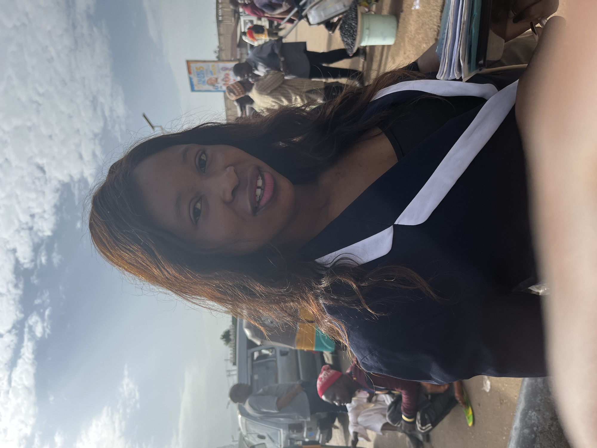 A woman with long, dark hair, wearing a dark blazer with white trim over a black top, is the primary subject in the foreground of this outdoor, daytime photograph. She is looking towards the camera with a slight smile. Behind her, in the mid-ground, is a busy street scene in Gura Top, Nigeria. Several people are visible, including men in traditional attire and others in more casual clothing. Vehicles, including vans and what appears to be a three-wheeled auto-rickshaw, are present. A market stall with a bucket of dark items, possibly produce or goods, is to the right. The background shows buildings and a bright, cloudy sky. The ground is paved with dirt and some debris. A small digital tablet or book is held in the woman's right hand, and she appears to be holding a pen.