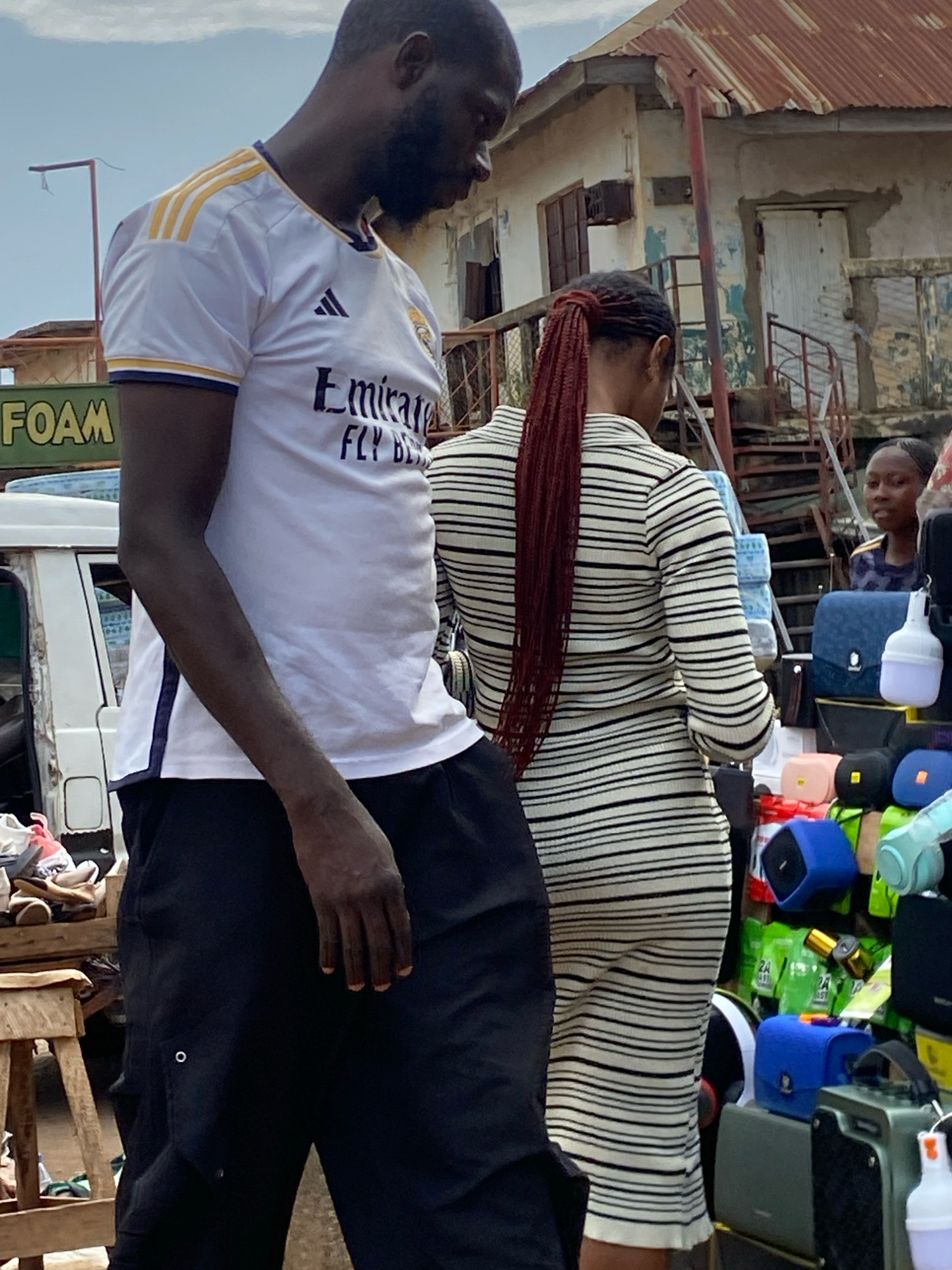 A man wearing a white t-shirt with yellow stripes on the shoulders and a black graphic that reads "Fly Better" stands facing away from the camera in Bukuru, Nigeria. He has dark skin and a beard, and is wearing black pants. To his right, a woman with dark skin and braided red hair, also facing away from the camera, wears a black and white striped dress. Behind them, a market stall is visible, displaying various bags and other items, including several blue and green speakers. A young woman is seen looking from behind the market stall. The environment appears to be an outdoor market with some buildings and a cloudy sky.