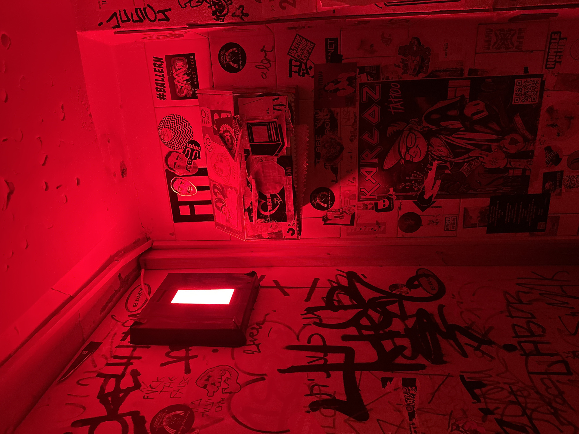 This image captures a vibrant, narrow indoor space, likely a hallway or passage, entirely bathed in an intense red light emanating from an unseen source and a visible rectangular light fixture on the lower left wall. The setting suggests an urban, underground, or alternative venue in Tel-Aviv, Israel, given the blend of Hebrew and English text.
The walls are densely covered with a layered collage of graffiti, stickers, and posters, creating a chaotic yet expressive aesthetic. Various pieces of graffiti in black spray paint and markers, including illegible tags, "FVCK," "LABOR," "HERS," and "FAL," are visible. Numerous stickers adorn the surfaces, some featuring stylized faces and abstract patterns. A 3D box-like structure, also covered in stickers, juts out from the upper wall.
Notable posters include a large one depicting a cartoon-like character, possibly a rabbit or similar, with glasses and a cap, next to the words "KARLAZE TATTOO" and a QR code. Other visible texts on stickers and posters include "#BALLERN," "BIWS," "GOOD CREU," "HENGEL DEVICES," "ULTRAS CHEMIC LEIPZIG," "HUMBLE CLUB," and "NET." Hebrew text such as "נעמה גיל" (Naama Gil), "מה קורה מיום" (What's up from day/since day), and "מך" are also present. The overall atmosphere conveyed by the red lighting and street art is one of an edgy, subcultural gathering place, potentially a bar, club, or art space. No specific activity is depicted, but the environment itself speaks of active participation and expression.