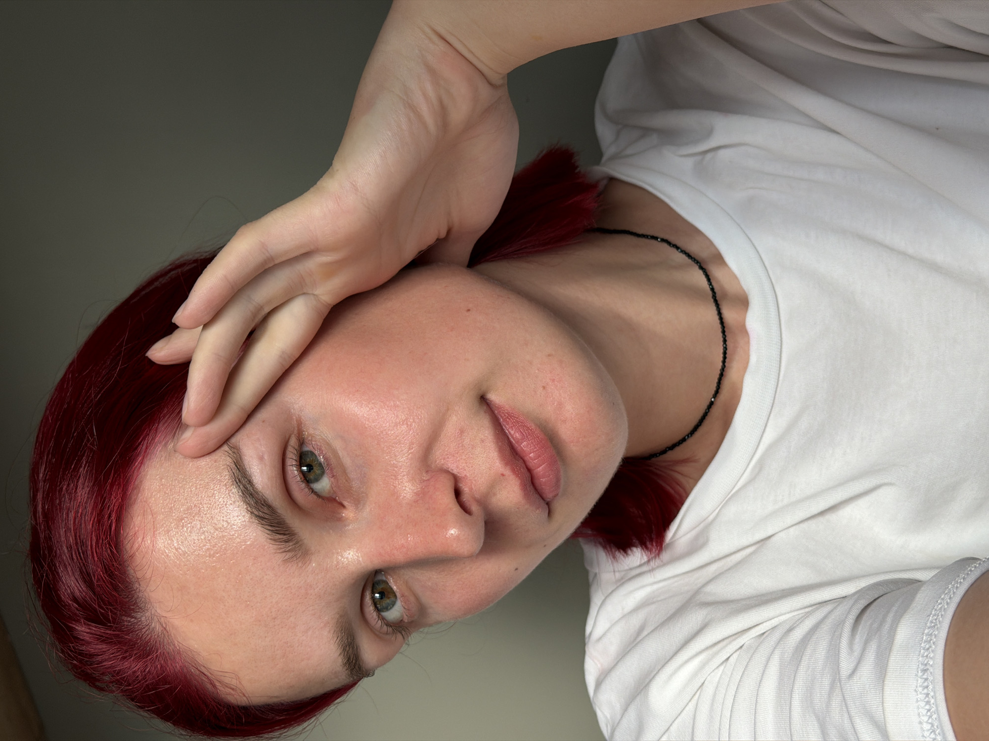This close-up portrait features a young individual, likely female, with vibrant red hair, fair skin, and light green or hazel eyes. They are wearing a plain white crew-neck t-shirt and a thin, black beaded necklace. The person looks directly into the camera with a calm, neutral expression. One hand is gently placed on their forehead, partially covering the temple area. The image appears to be a selfie, taken at a slight upward angle. The background is a simple, plain light grey or off-white wall, suggesting an indoor setting. The lighting is soft and even, without harsh shadows. No specific location cues are visible within the image to indicate Chaikovsky, Russia, nor is any text visible. The time of day and weather are not discernible.