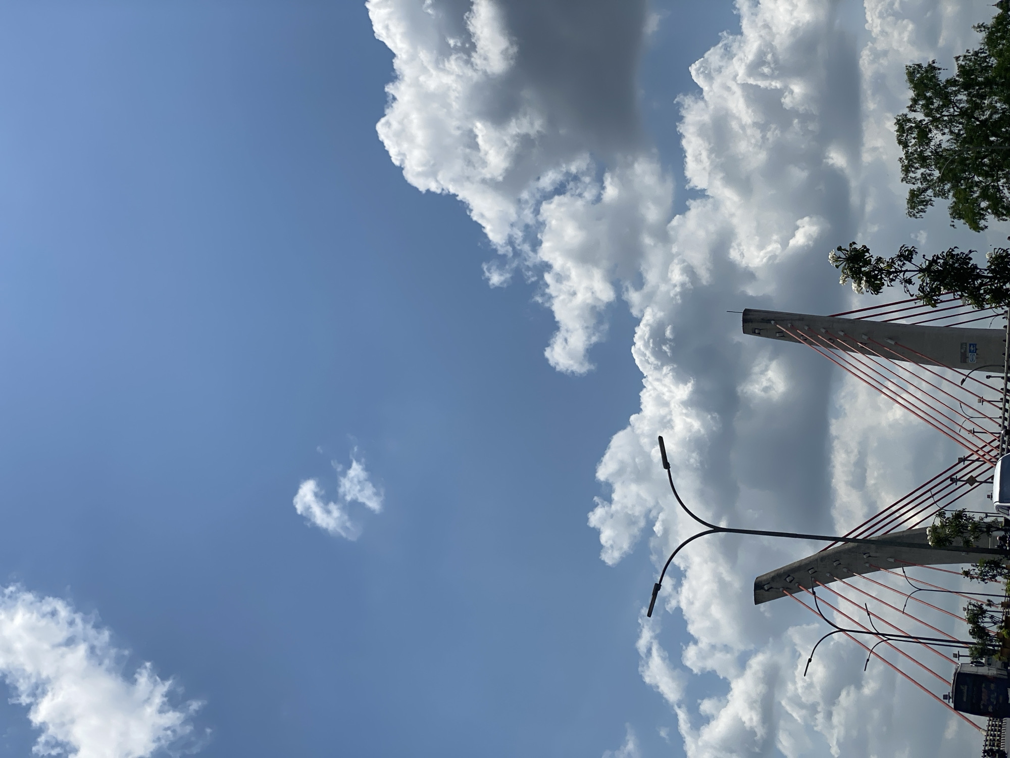 The image captures a daytime view of a bridge with distinctive concrete towers and red suspension cables, located in Banjarmasin, Indonesia. The sky is partly cloudy, with large, puffy white and gray clouds dominating the upper portion of the frame, set against a clear blue sky. The bridge's structure, including streetlights attached to curved poles, is visible in the lower half of the image. Vegetation, including trees and smaller plants, lines the roadway beneath the bridge. The scene is observed from a low angle, looking upwards towards the bridge and the sky, suggesting the photograph was taken from ground level or a nearby vehicle. The presence of vehicles, including a bus and a car, indicates that the bridge is in active use. The overall impression is one of an urban infrastructure element integrated into a natural setting, under a dynamic sky.