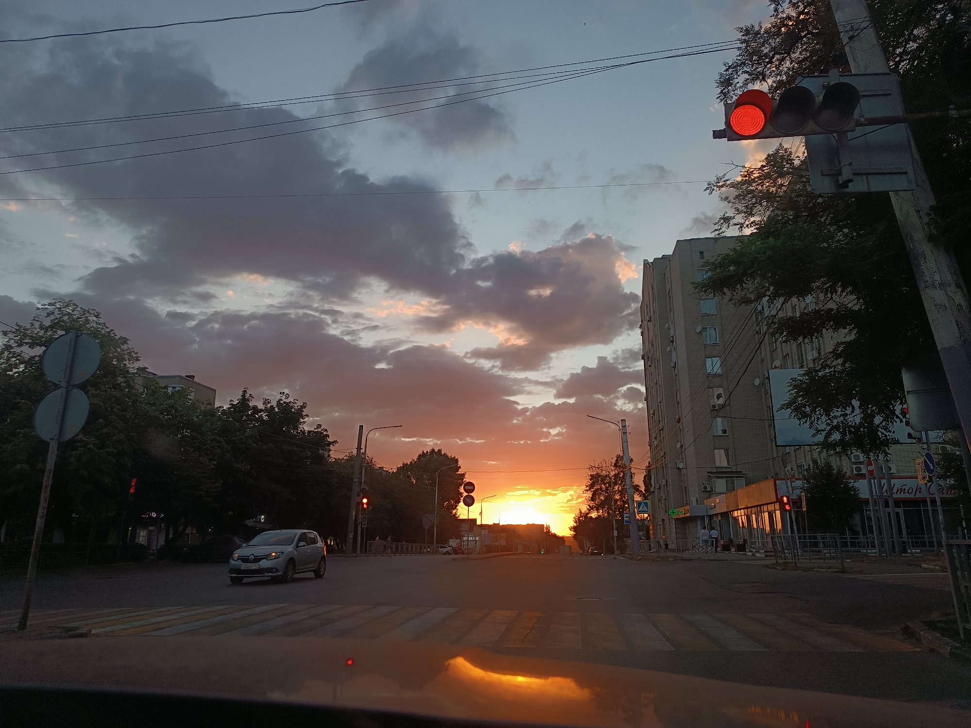 An urban intersection is depicted at sunset. The sky is filled with dramatic clouds, with a vibrant orange glow emanating from the setting sun. A multi-story building stands on the right side of the frame, its facade reflecting the warm light. On the left, trees line the street and a traffic light shows a red signal. A pedestrian crossing is visible in the foreground. A silver car is driving away from the camera, and another red traffic light is visible in the distance. The reflection of the sky and a traffic light is seen on the wet surface in the foreground.