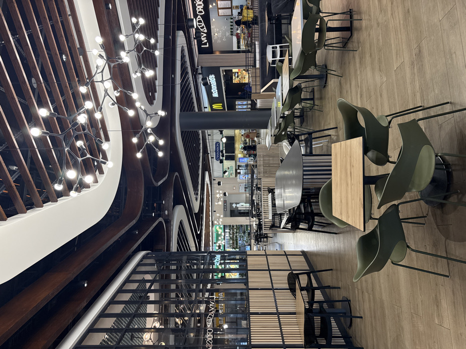 This image depicts the interior of a large, modern food court, identified as being within the Blockbuster Mall in Kyiv, Ukraine. The high ceiling features a complex design with dark wooden slat panels interspersed with curvilinear white soffits, from which multiple contemporary chandeliers with black, branch-like structures holding numerous spherical light bulbs are suspended. The floor is composed of light brown wood-effect tiles.
In the foreground and mid-ground, numerous clusters of dining tables and chairs are arranged. Tables vary in shape, including square, rectangular, and elongated oval, with tops of light wood laminate or dark gray material, supported by dark metal bases. The chairs are primarily dark olive green or black plastic, with some white chairs, all featuring modern designs and dark metal legs. The majority of these seating areas are unoccupied.
In the background, a row of food establishments is visible. Prominent signs identify "Lviv Croissant" (right) and "McDonald's" (center-right). The sign for "Cinnabon" is also discernible to the left of McDonald's. Few individuals are present; one person in a light-colored coat walks in the mid-ground near Cinnabon, and several others are vaguely visible standing or moving near the counters of Lviv Croissant and McDonald's. Dark gray cylindrical pillars support the ceiling structure. Sections of the food court are delineated by partitions featuring vertical light wood slats set within dark metal frames.