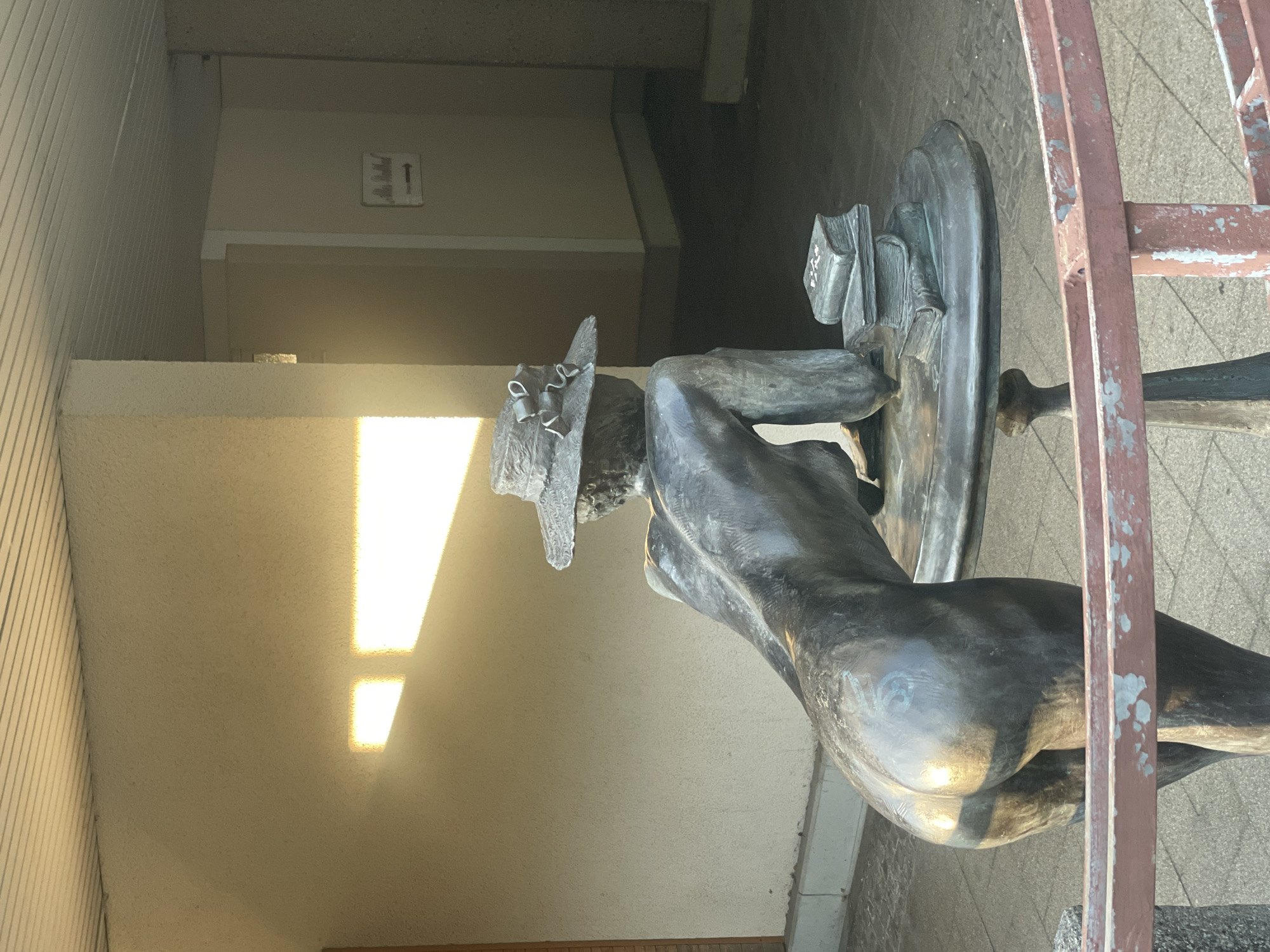 This image shows a bronze sculpture of a nude woman wearing a hat, bent over a table with a stack of books. The sculpture is located in front of a building with textured walls and a slatted ceiling. Sunlight streams in from the left, casting a rectangle of light on the wall. A sign with an arrow is visible on a wall in the background. The foreground features a reddish-brown metal railing. The location is the 4. Mai-Platz in Iserlohn, Germany, specifically the sculpture "Lesende" (The Reader) by Ursula Strigel.