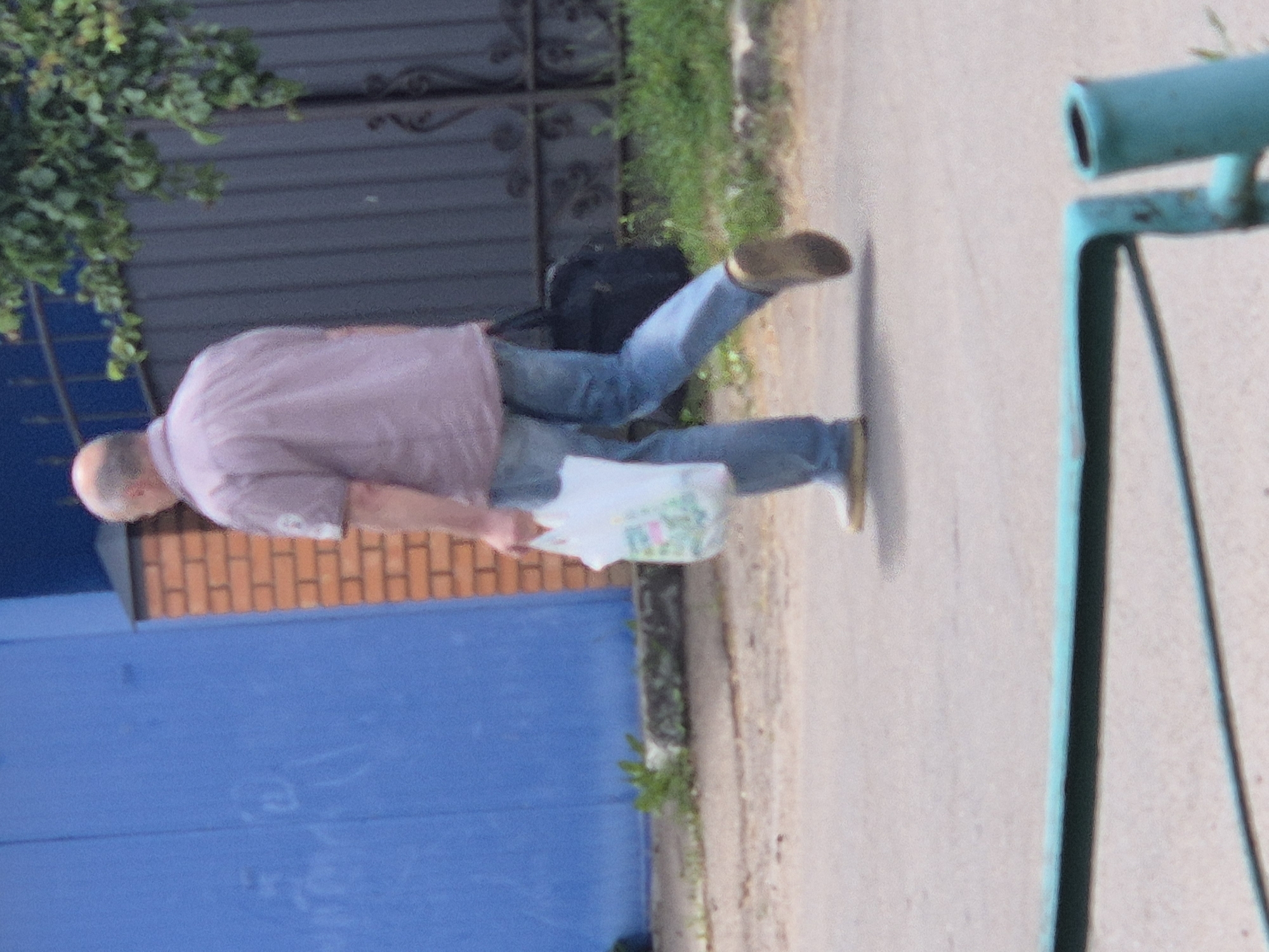 A middle-aged, bald man with fair skin is walking away from the camera on a paved surface in Sozonivka, Ukraine. He is wearing a light purple, short-sleeved button-up shirt and blue jeans. In his right hand, he is carrying a white plastic shopping bag with colorful images of food items. A dark-colored duffel bag is slung over his left shoulder. The background features a blue metal door or wall on the left, a brick structure behind the man, and a black metal fence with decorative elements to the right, partially obscured by green foliage from a tree. The ground is a mix of pavement and dirt. A turquoise metal railing is visible in the foreground.