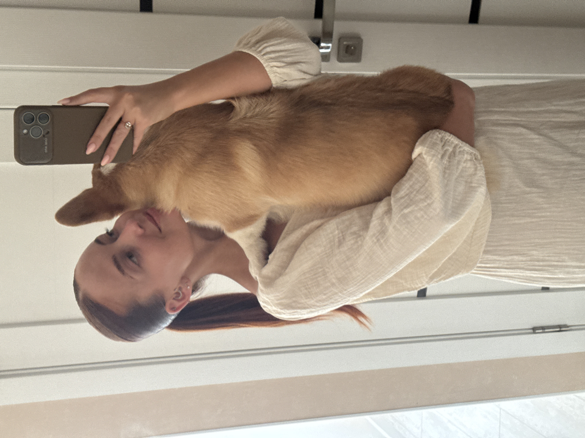 A person is holding a corgi dog while taking a selfie with a smartphone. The individual is wearing a light-colored, loose-fitting top. The background appears to be a room with white walls and a light-colored floor. The location is not specified as Chaikovsky, Russia, in the image.