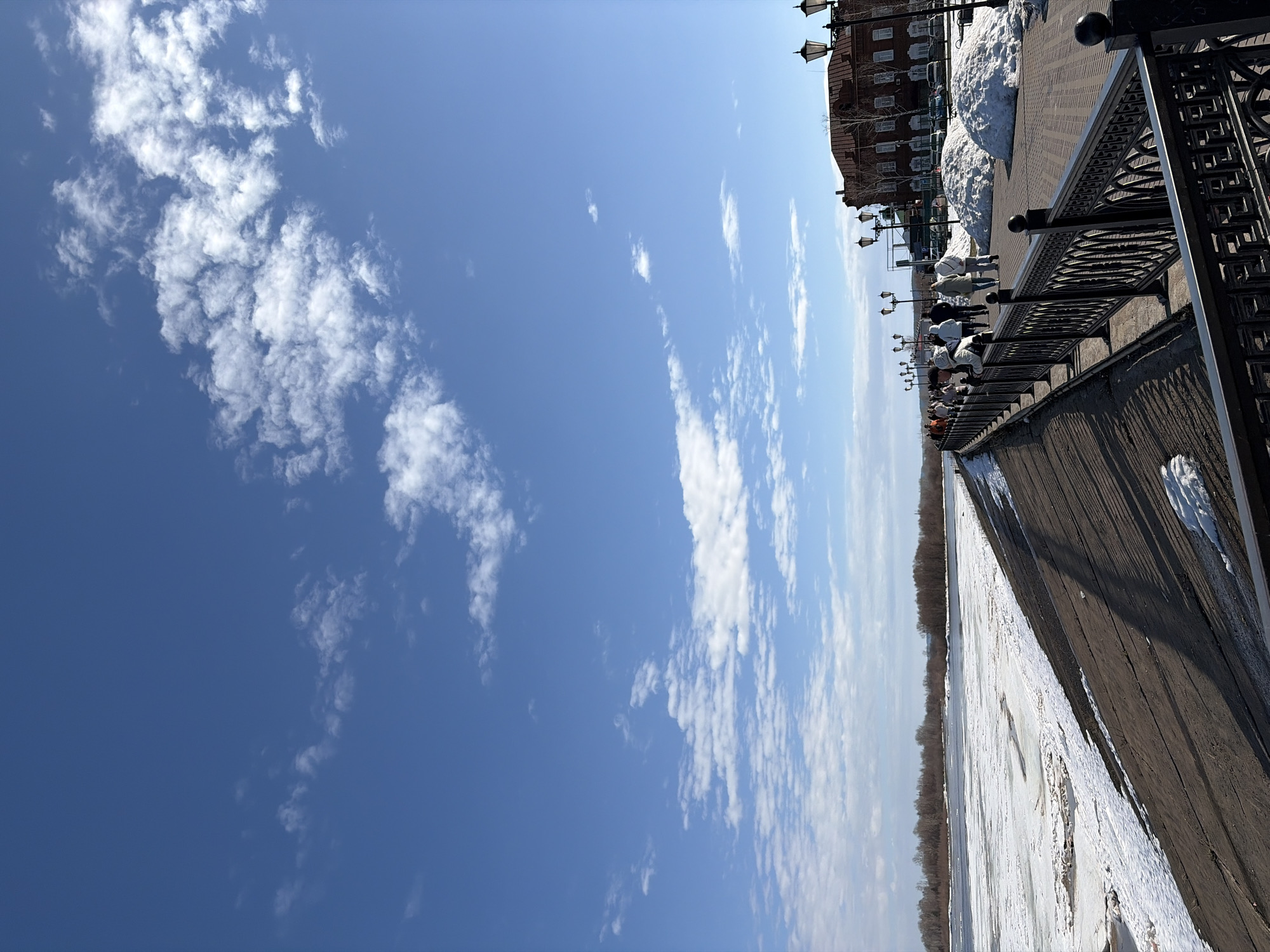 The image depicts a view from the embankment in Sarapul, Russia, overlooking the Kama River. The river is partially frozen and covered with snow. The embankment features a wrought iron railing, a paved walkway, and a row of streetlights. Several people are visible along the embankment, seemingly enjoying the view. In the background, a brick building with multiple windows and snow piled near its base can be seen. The sky is blue with scattered clouds. The orientation of the image is rotated clockwise by 90 degrees.