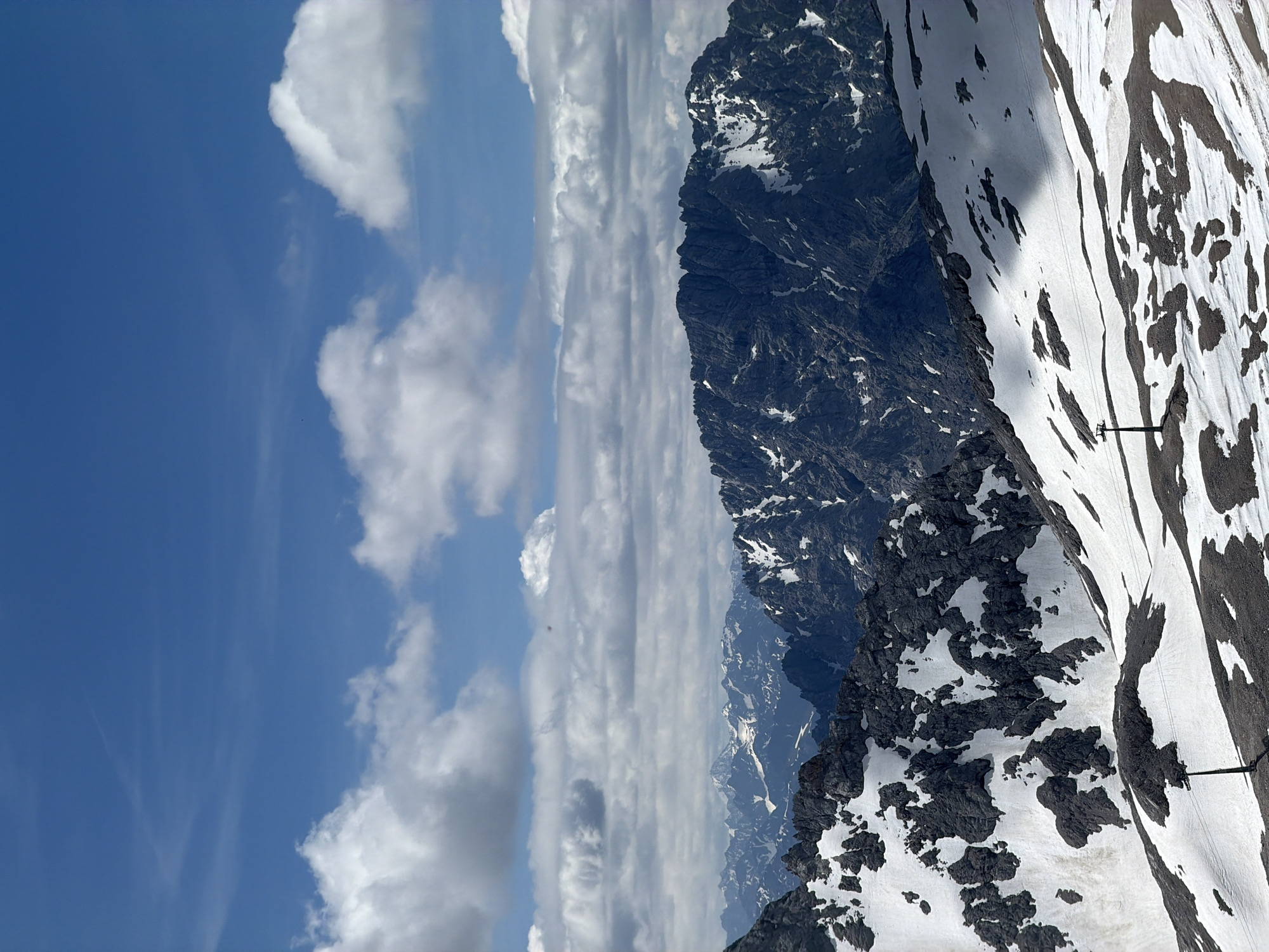 This is a landscape photograph taken from a high vantage point, likely in the Bavarian Alps near Garmisch-Partenkirchen, Germany. The scene depicts a dramatic mountainous terrain under a partly cloudy blue sky. In the foreground, a snow-covered slope with patches of exposed dark rock leads the eye towards rugged, dark gray mountain peaks in the midground and background. These mountains are also partially covered in snow, highlighting their jagged contours. The sky above is a vibrant blue, populated by scattered white cumulus clouds, some appearing puffy and others more wispy. One of the clues pointing to this location is the presence of a ski lift tower on the snowy slope in the foreground, suggesting a ski resort or a mountain accessible by such infrastructure, which is common in the Garmisch-Partenkirchen area. The overall impression is one of a vast, natural, and somewhat stark alpine environment.