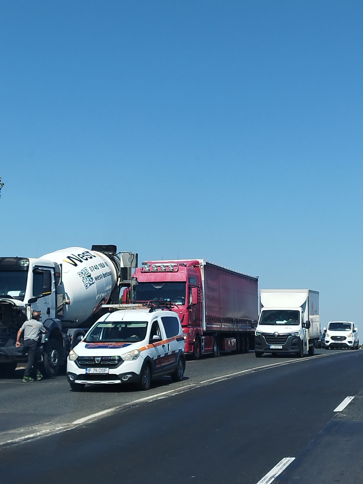 In Chitila, Romania, on a sunny day with a clear blue sky, a traffic incident has occurred on a multi-lane road. A white cement mixer truck with "West" branding is parked on the left side of the road, partially blocking a lane. To its right, a red semi-trailer truck with a long red trailer is also stopped. In front of these larger vehicles, a white and orange service van, possibly an emergency vehicle, is positioned. Further down the road, a white panel van and a white utility vehicle are also stopped in traffic. A person wearing a grey shirt and dark pants is standing near the cement mixer truck. The road surface is asphalt with clear white lane markings.